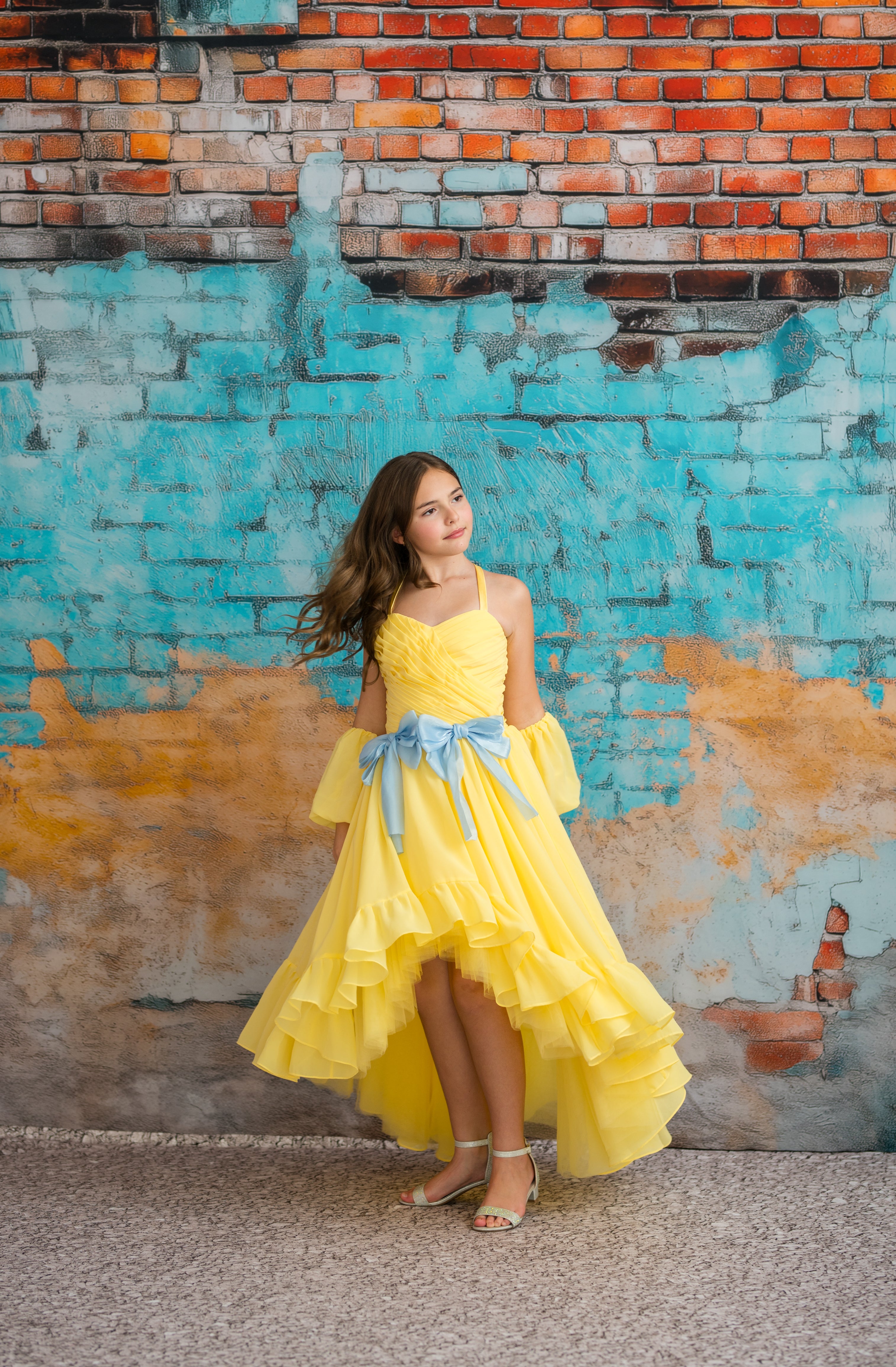 Woman in a yellow dress standing against a colorful brick wall.