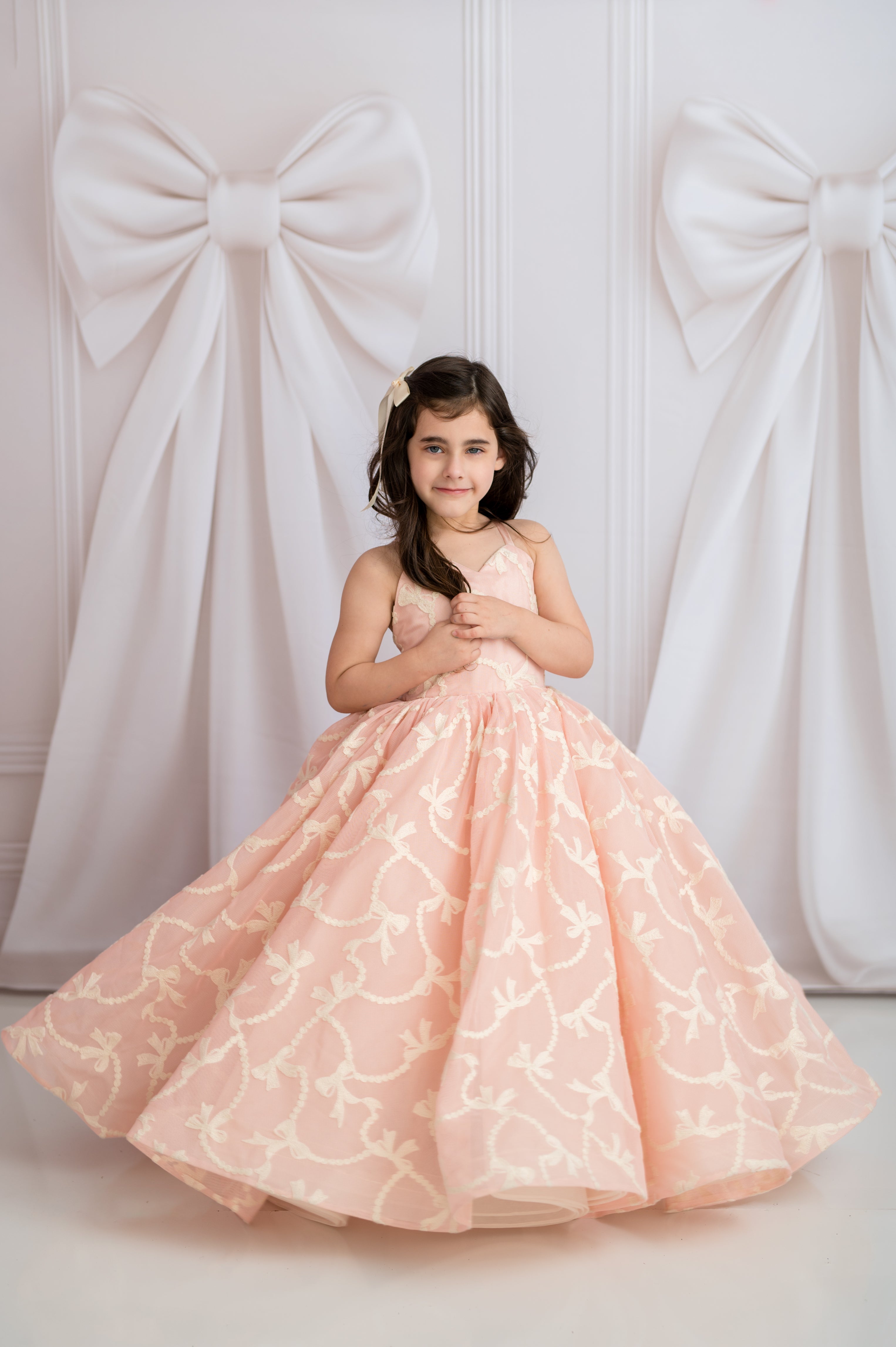 Young girl in a pink floral dress standing against a white wall with large decorative bows.