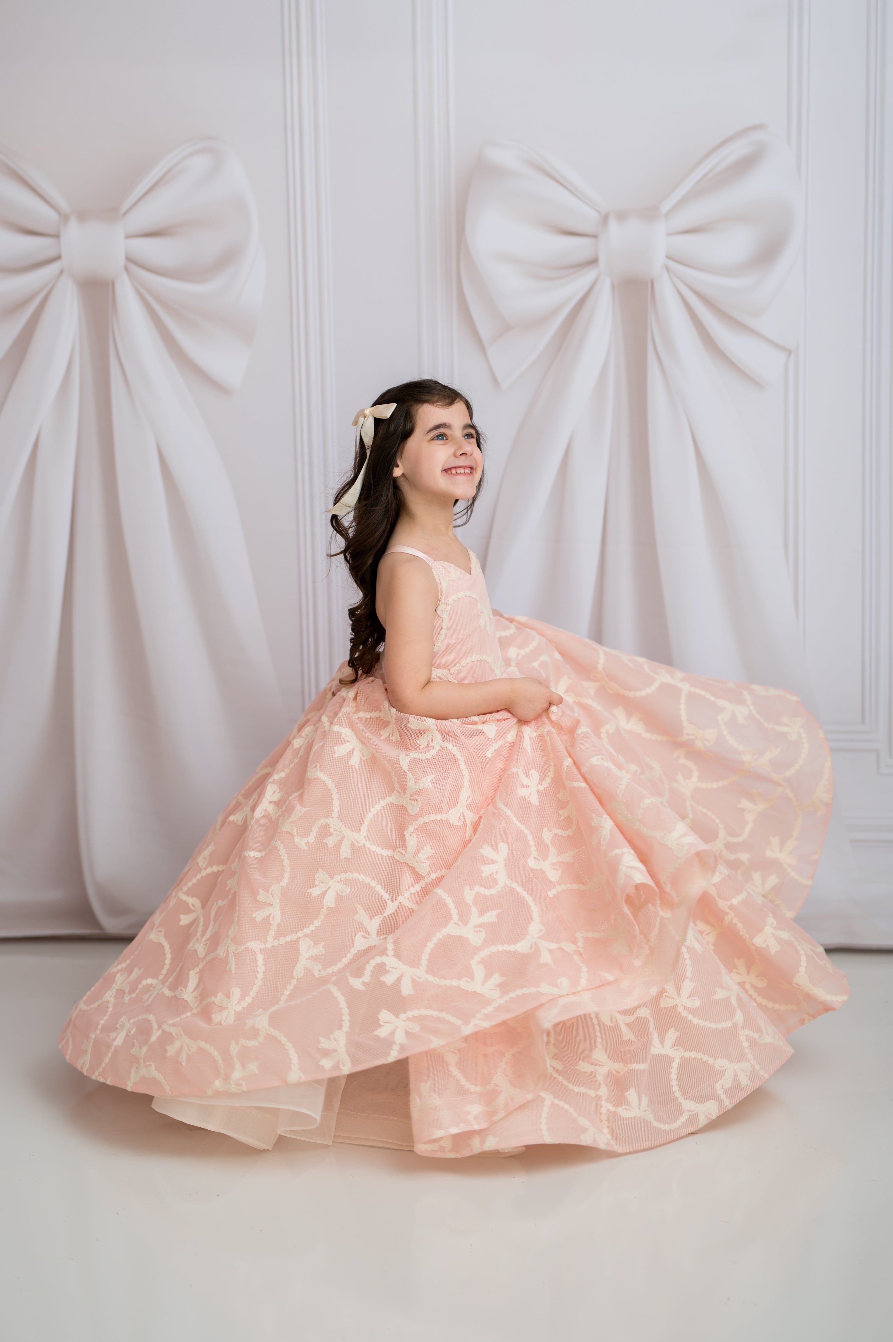 Young girl in a pink floral dress sitting against a white wall with large fabric bows.