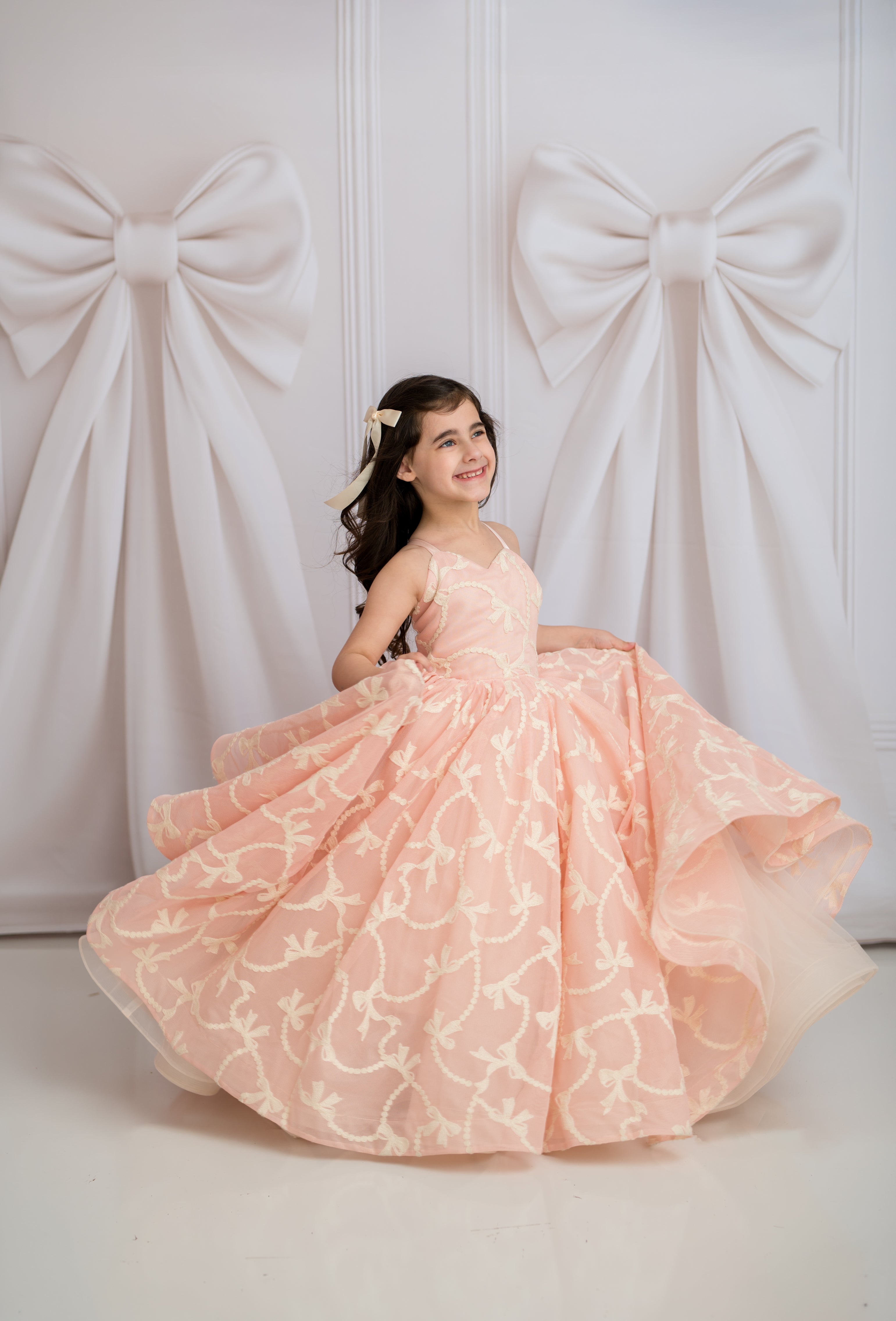 Young girl in a pink gown standing against a white wall with large decorative bows.