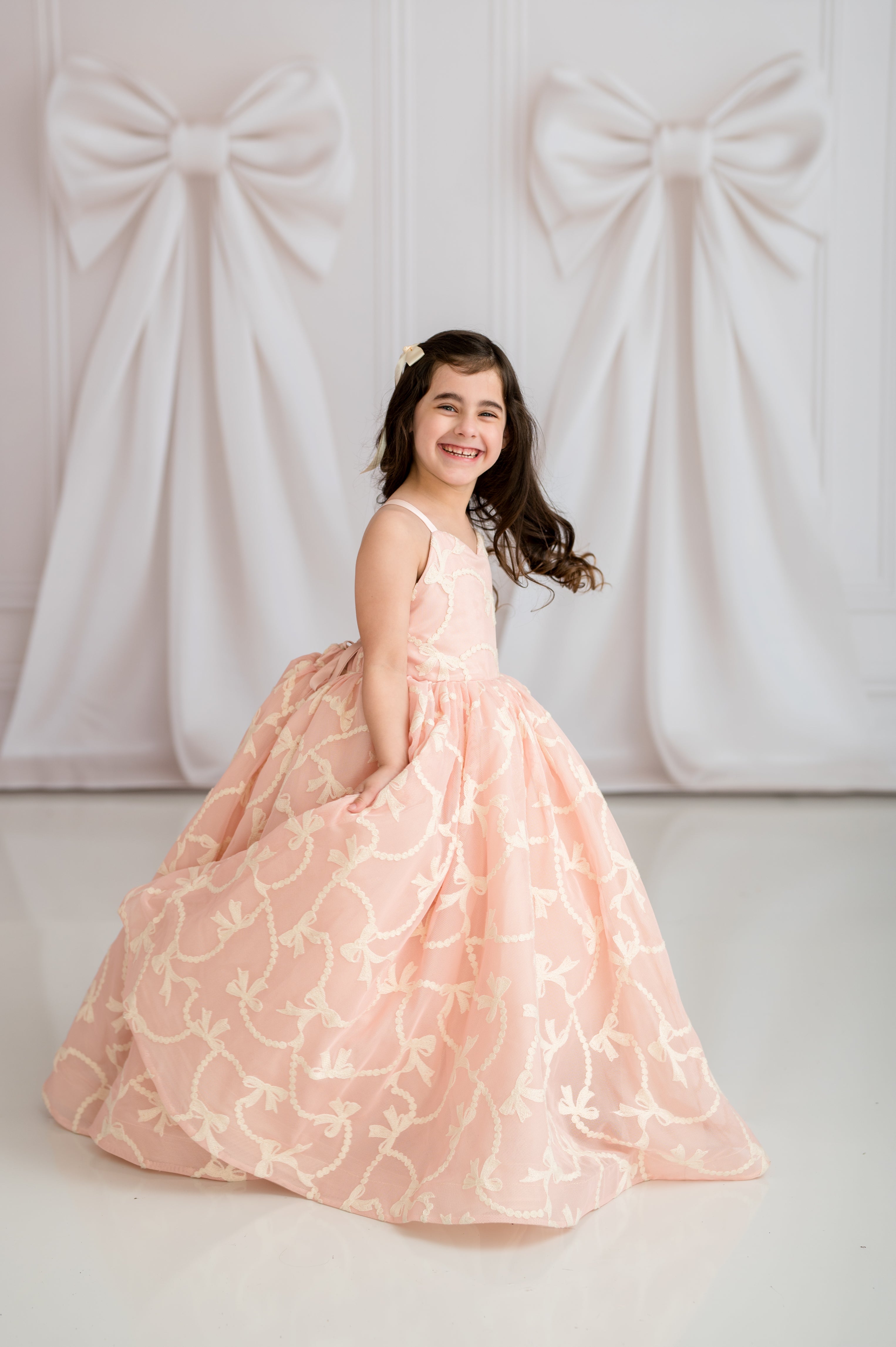 Young girl in a pink floral dress standing against a white background with large decorative bows.
