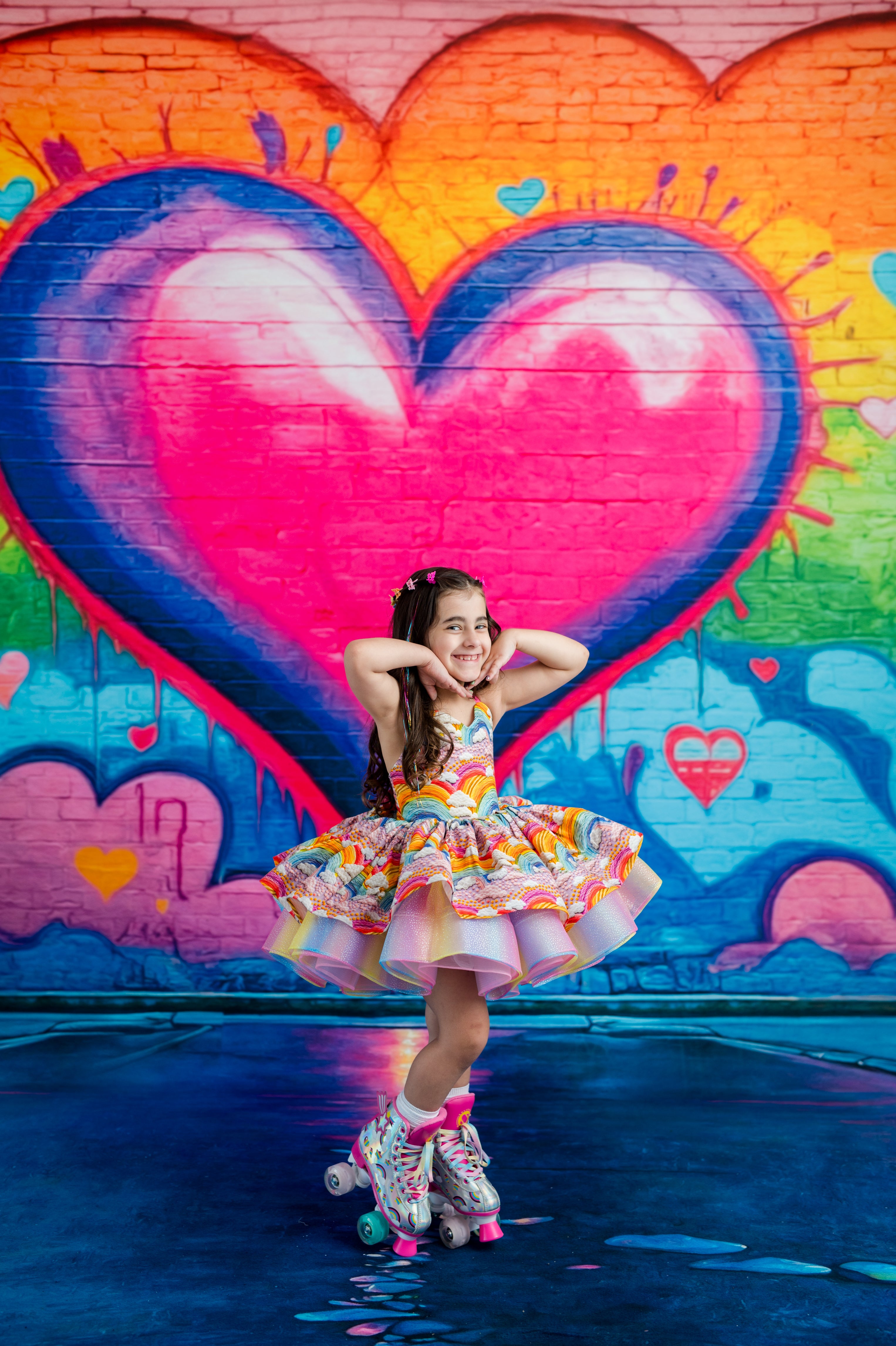 A young girl with long brown hair wearing a colorful ruffled tutu dress and roller skates poses in front of a vibrant graffiti wall featuring a large pink heart.