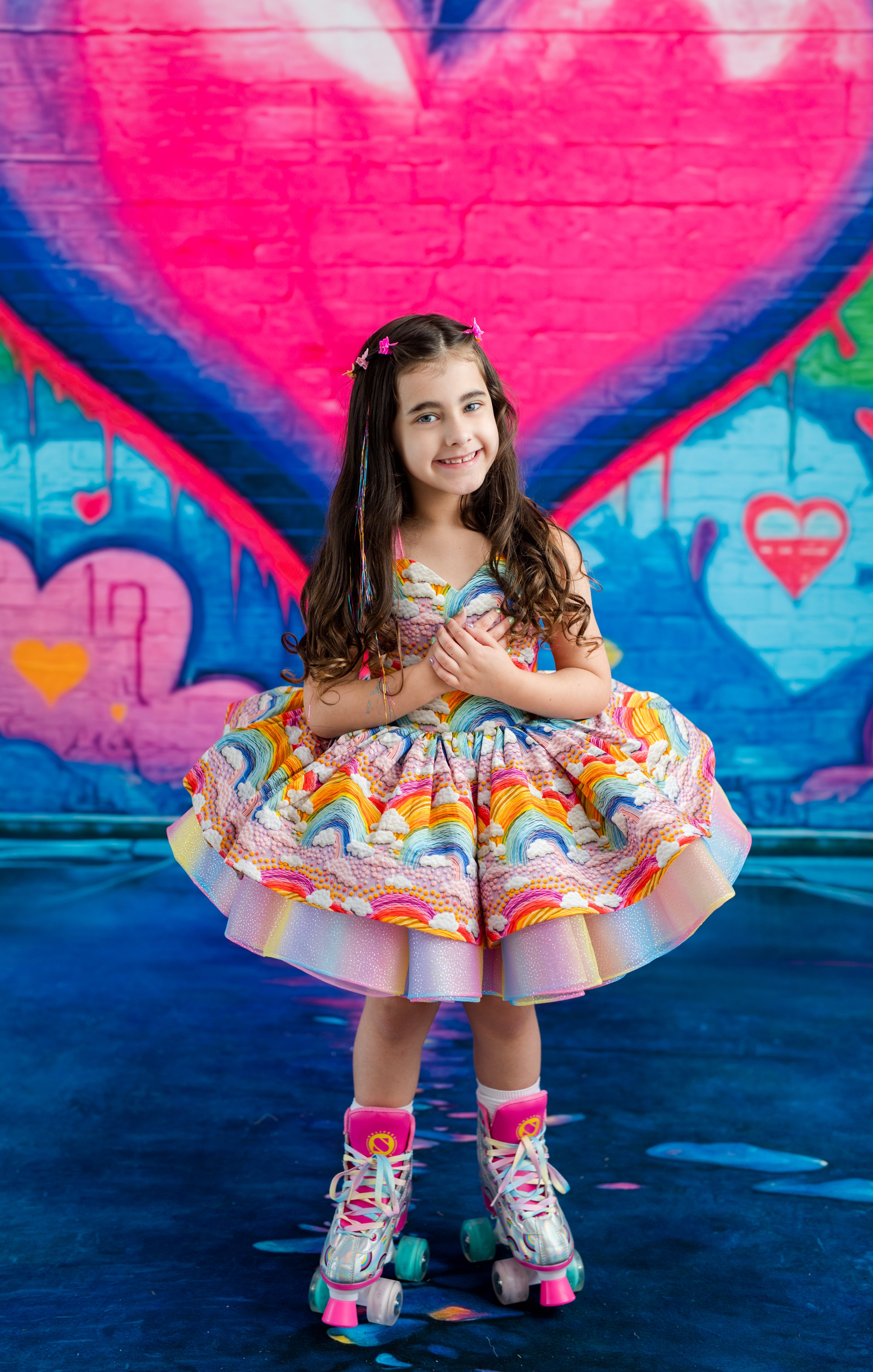 A young girl with long brown hair stands on roller skates in front of a colorful graffiti wall featuring a large pink heart. She is wearing a vibrant, multicolored ruffled dress with rainbow patterns.