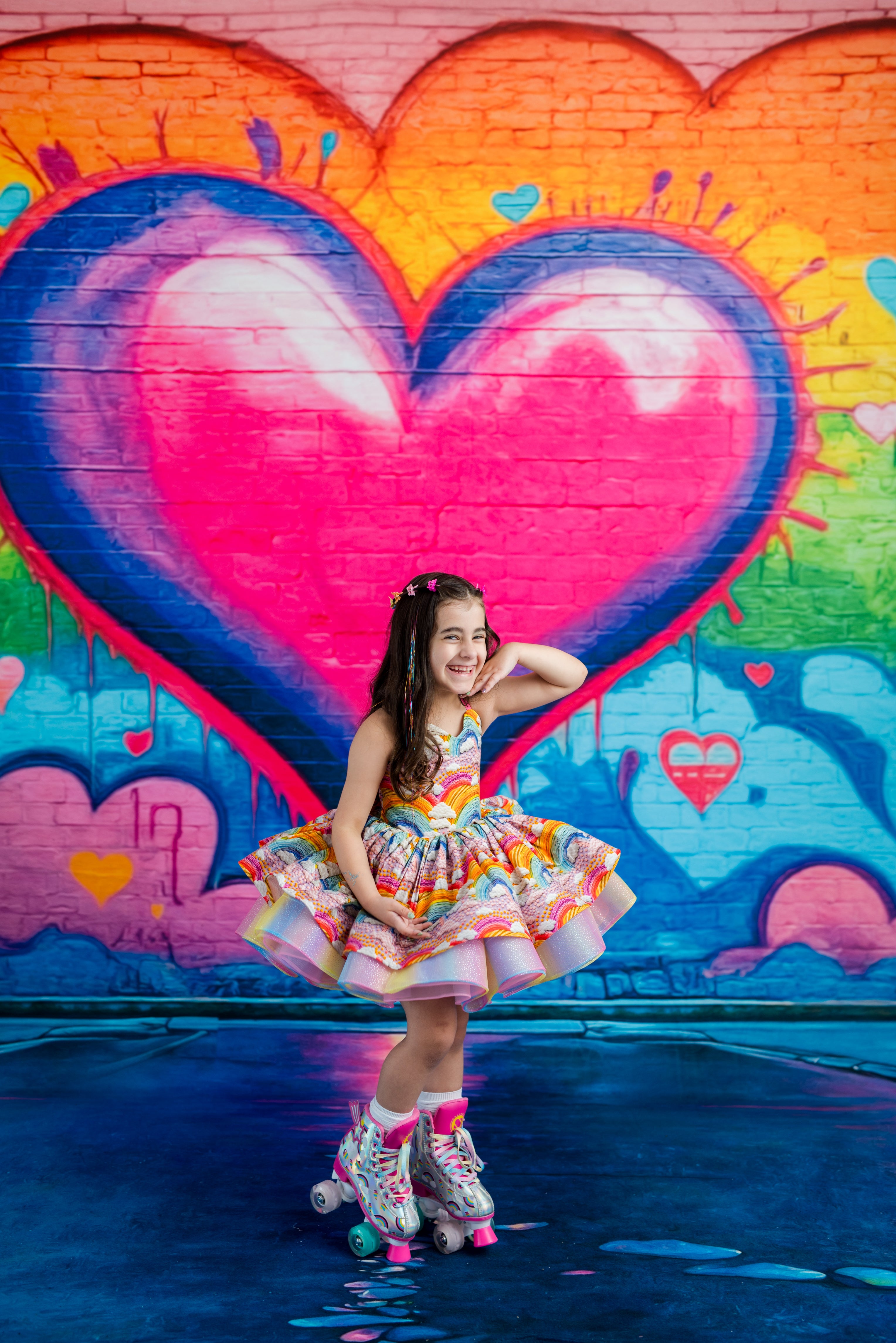 A young girl with long brown hair wearing a colorful floral print dress and roller skates poses in front of a vibrant graffiti wall featuring a large pink heart.