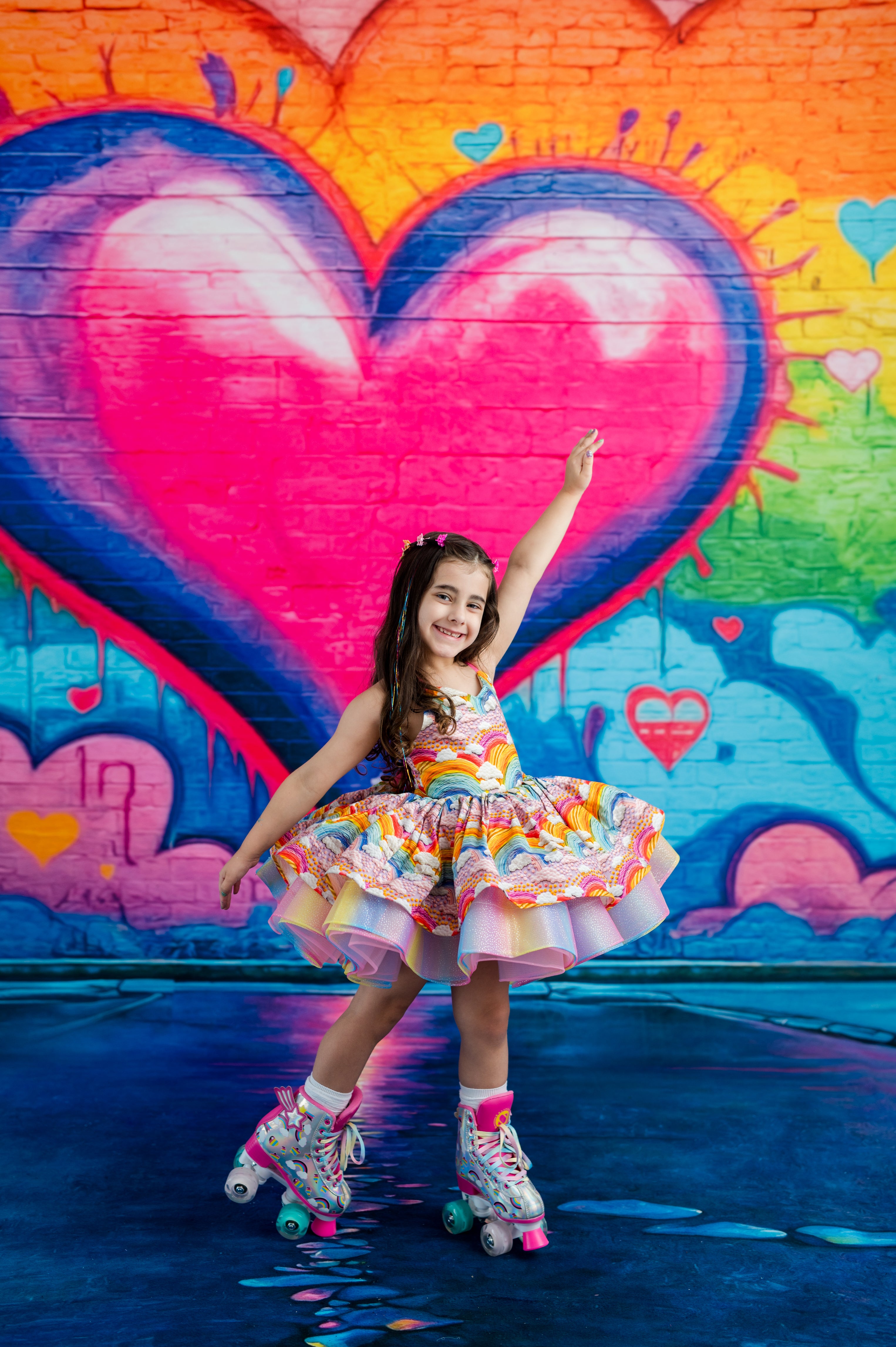 A young girl with long dark hair posing on roller skates in front of a vibrant graffiti wall featuring a large pink heart. She is wearing a colorful, tiered ruffled dress and matching roller skates.