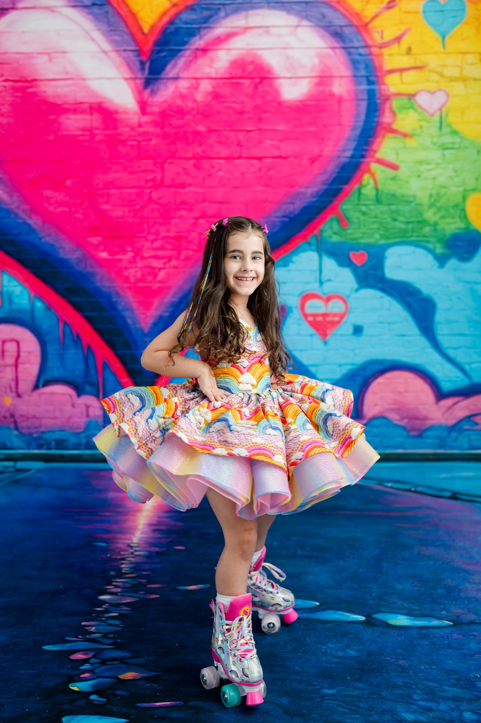 A young girl with long brown hair stands on roller skates in front of a colorful graffiti wall featuring a large pink heart. She is wearing a vibrant, multicolored ruffled dress with a white bodice and a bright, patterned skirt.