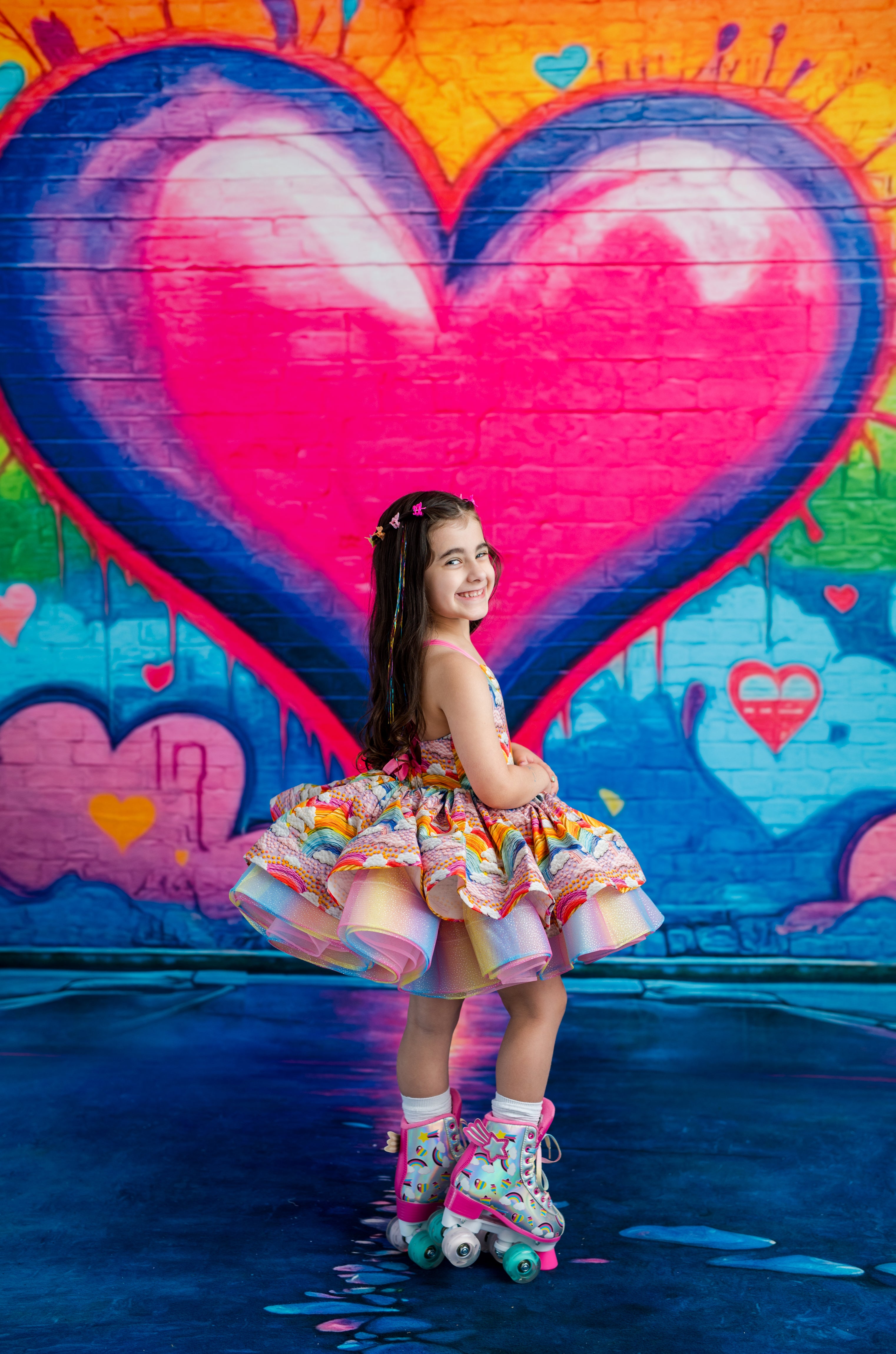 A young girl with long dark hair stands on roller skates in front of a vibrant graffiti wall featuring a large pink and blue heart. She is wearing a colorful tiered ruffle dress with pastel stripes and multicolor accents.