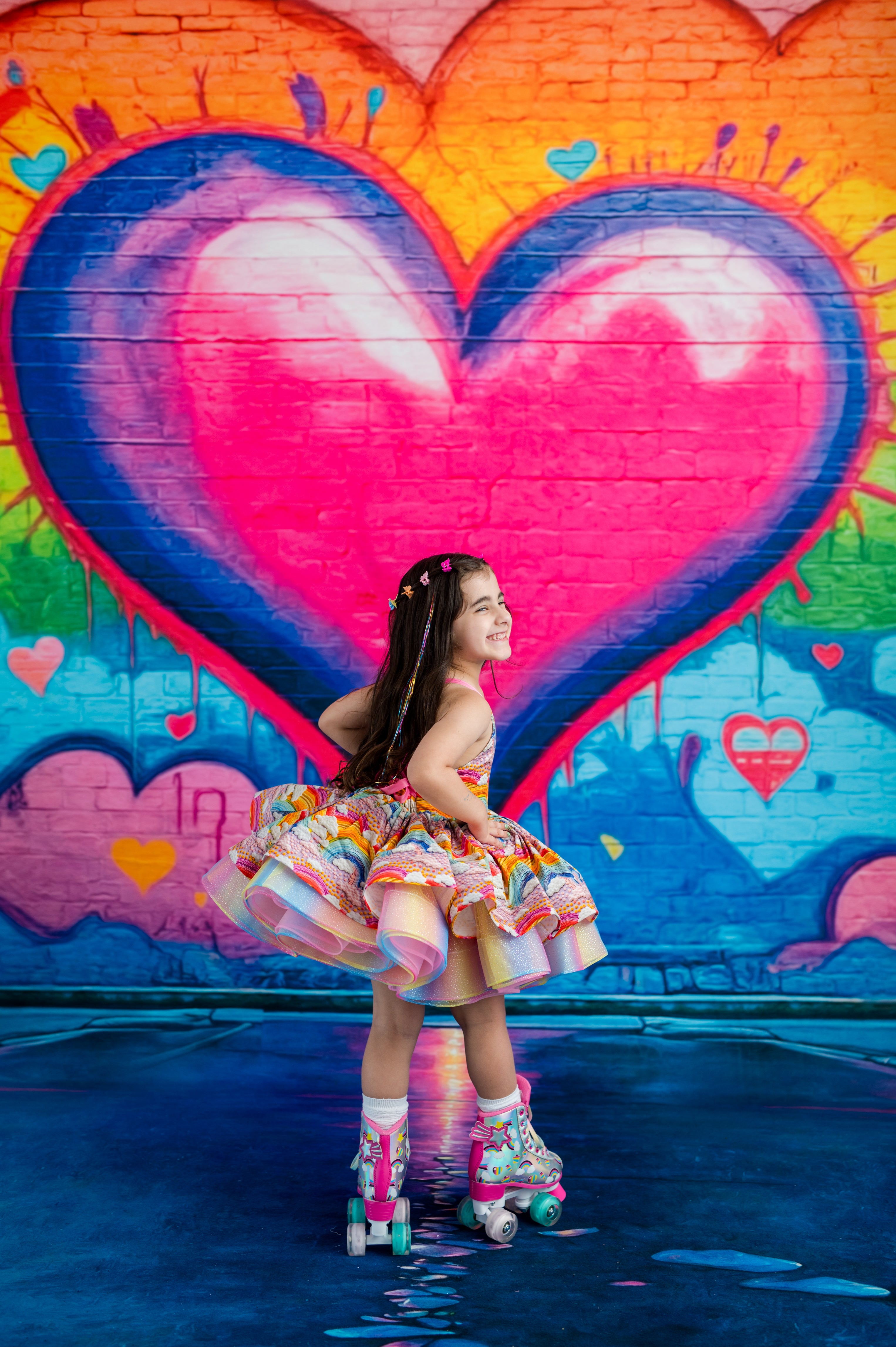 A young girl with long dark hair wearing a colorful floral tutu dress and roller skates poses in front of a vibrant graffiti wall featuring a large pink and blue heart.