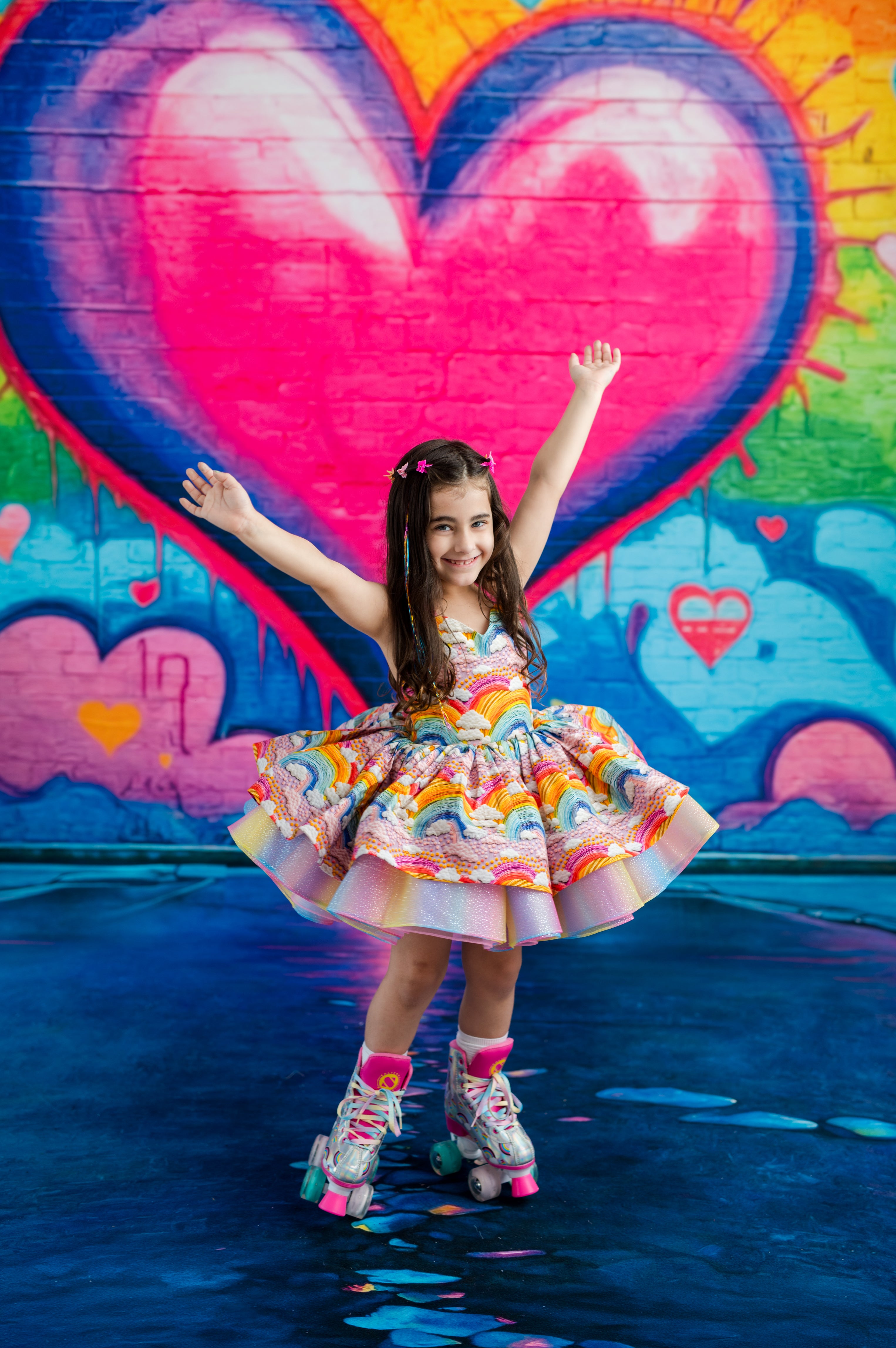 A young girl with long brown hair wearing a colorful rainbow-patterned tutu dress and roller skates poses on a wet blue surface in front of a vibrant graffiti wall featuring a large pink heart.