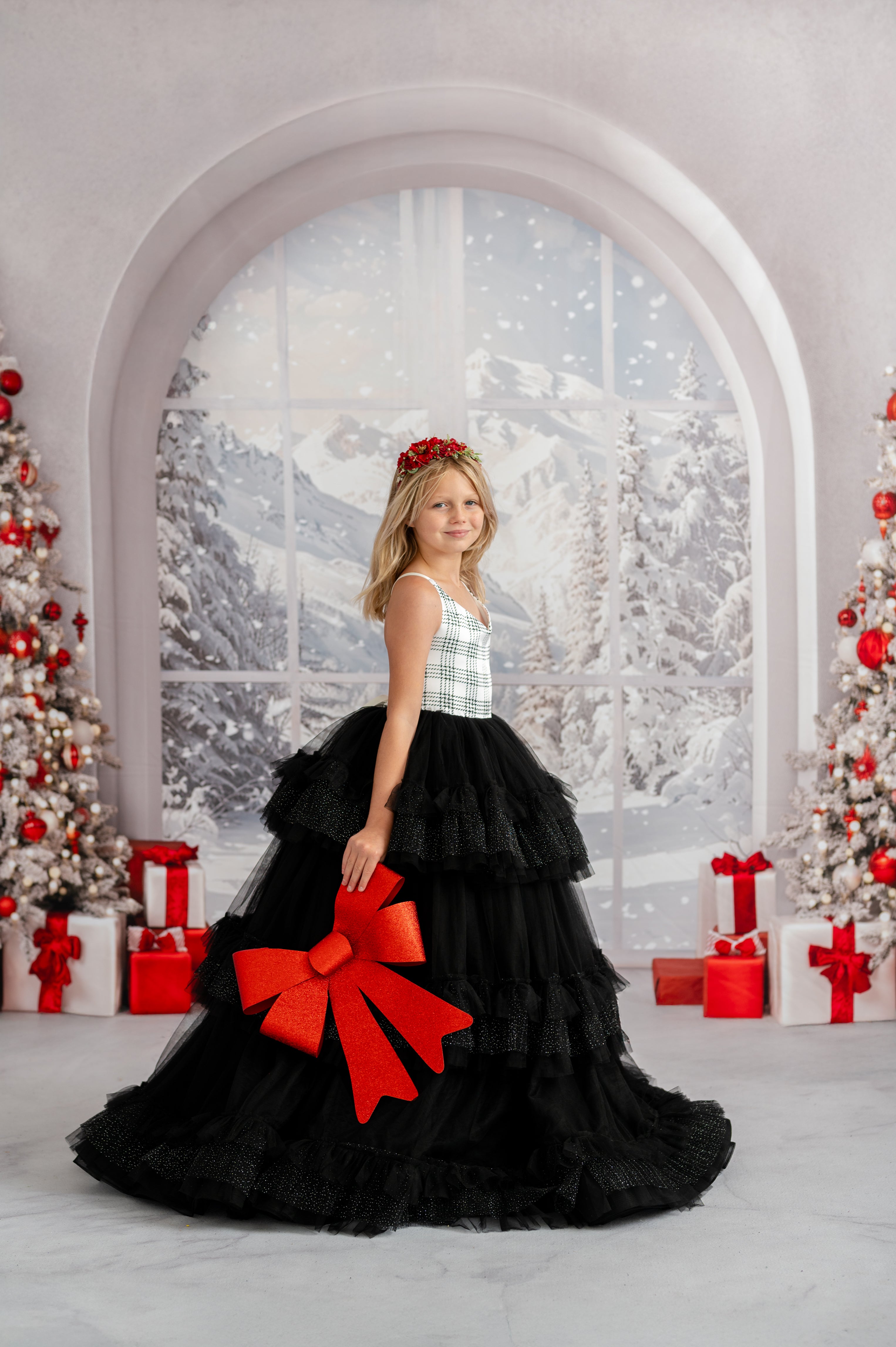 Young girl in a black dress with a large red bow standing in a festive setting with Christmas trees and presents.