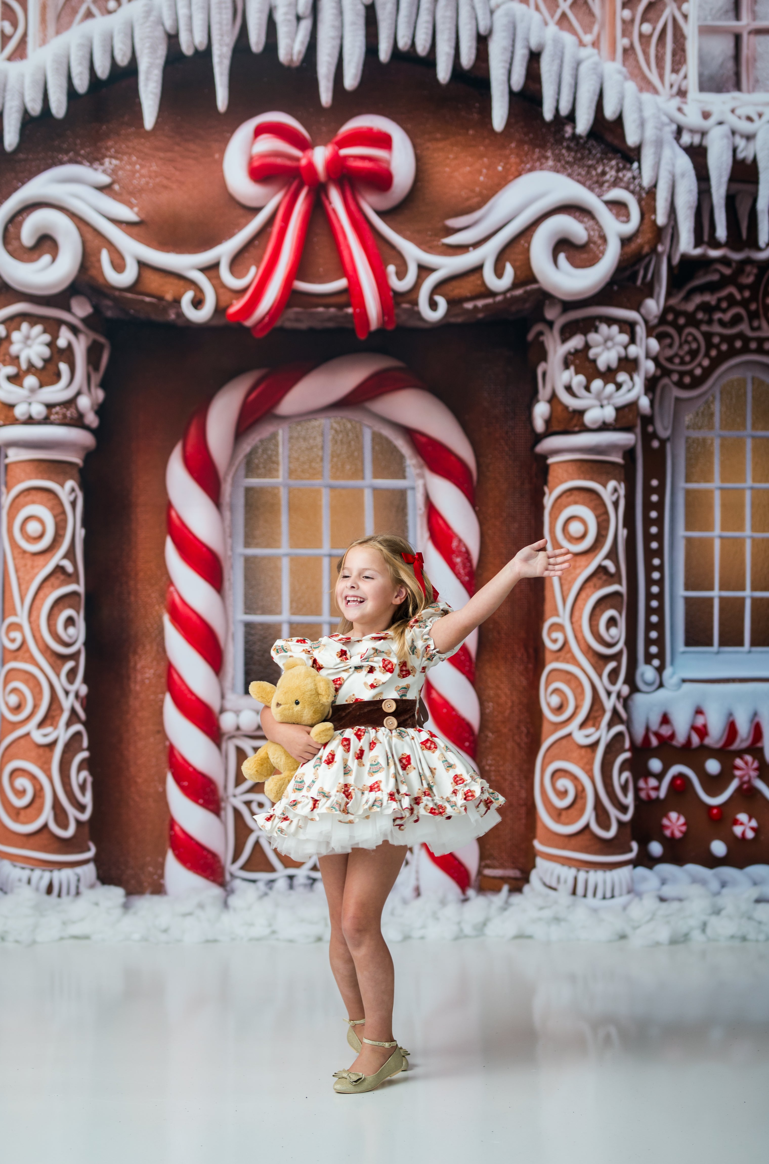 A young girl stands in front of a gingerbread house backdrop, wearing a white and red floral print dress with a brown belt and holding a yellow teddy bear.