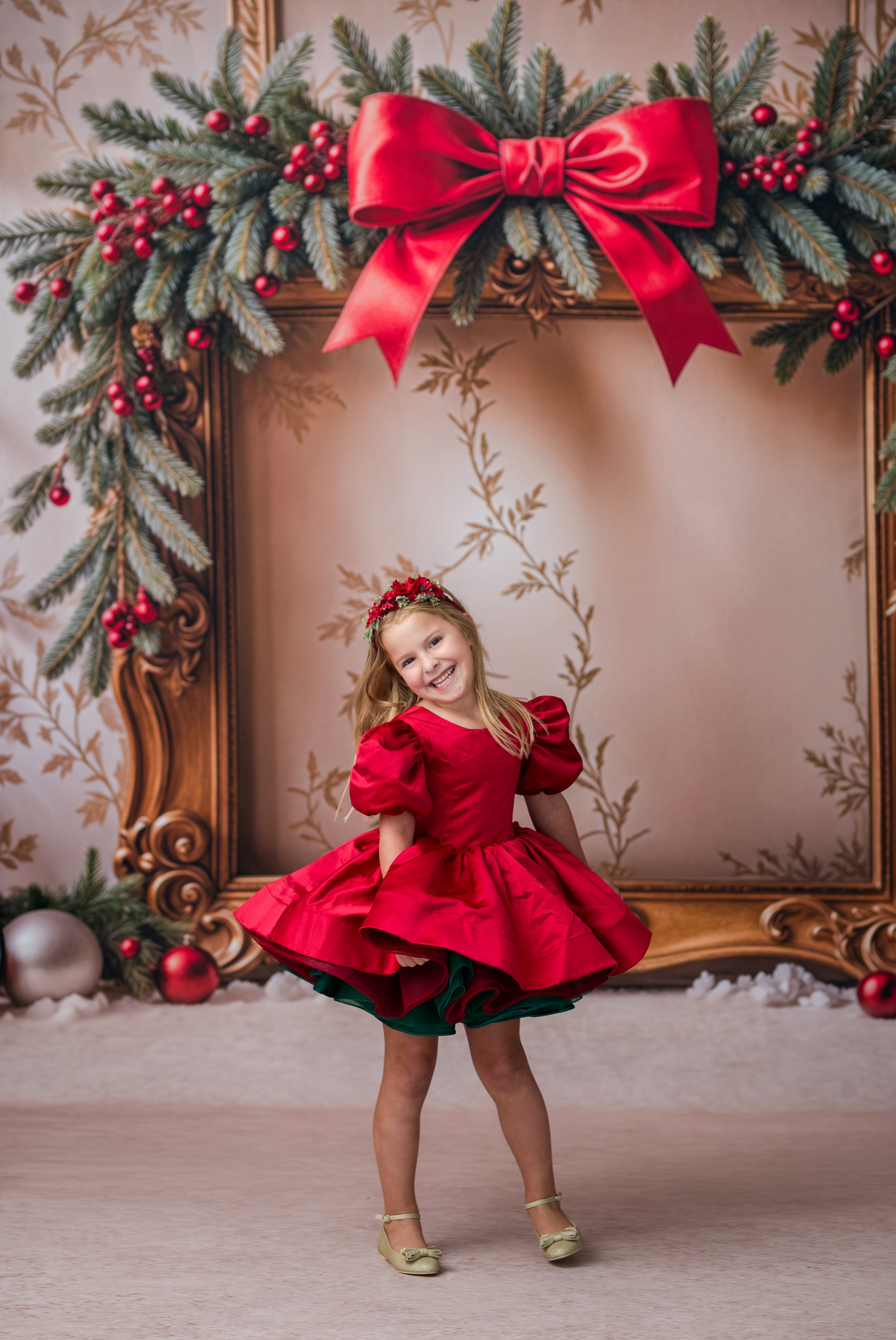 A young girl in a festive red and green holiday dress stands in front of an ornate gold picture frame decorated with a large red bow, pine branches, and red berries.