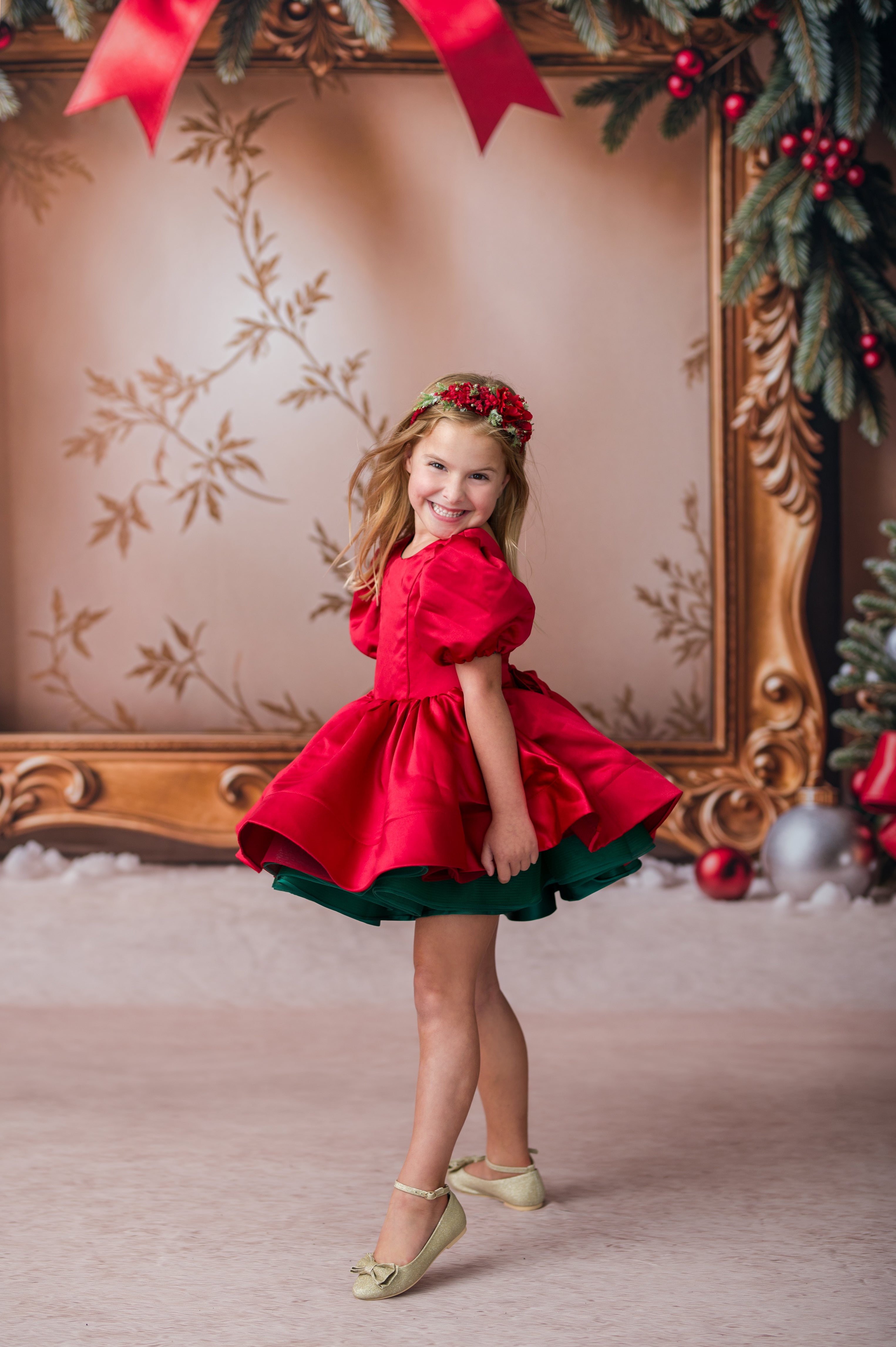 A young girl wearing a festive red and green holiday dress with a matching red flower crown, posing in front of an ornate gold picture frame decorated with Christmas greenery and red ribbons.