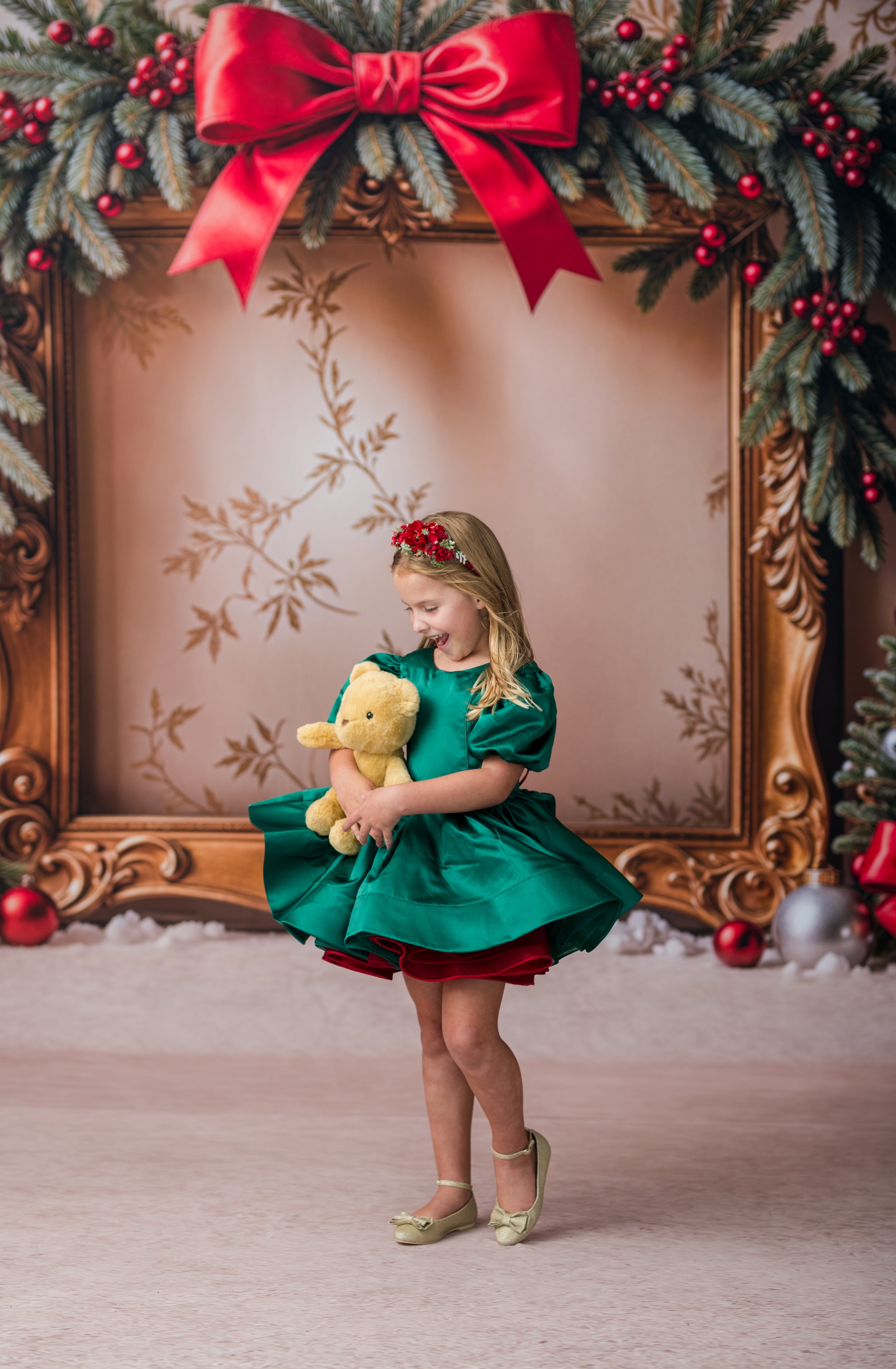 A young girl in a festive green and red holiday dress stands holding a yellow teddy bear in front of an ornate gold picture frame decorated with a large red bow and pine garlands.