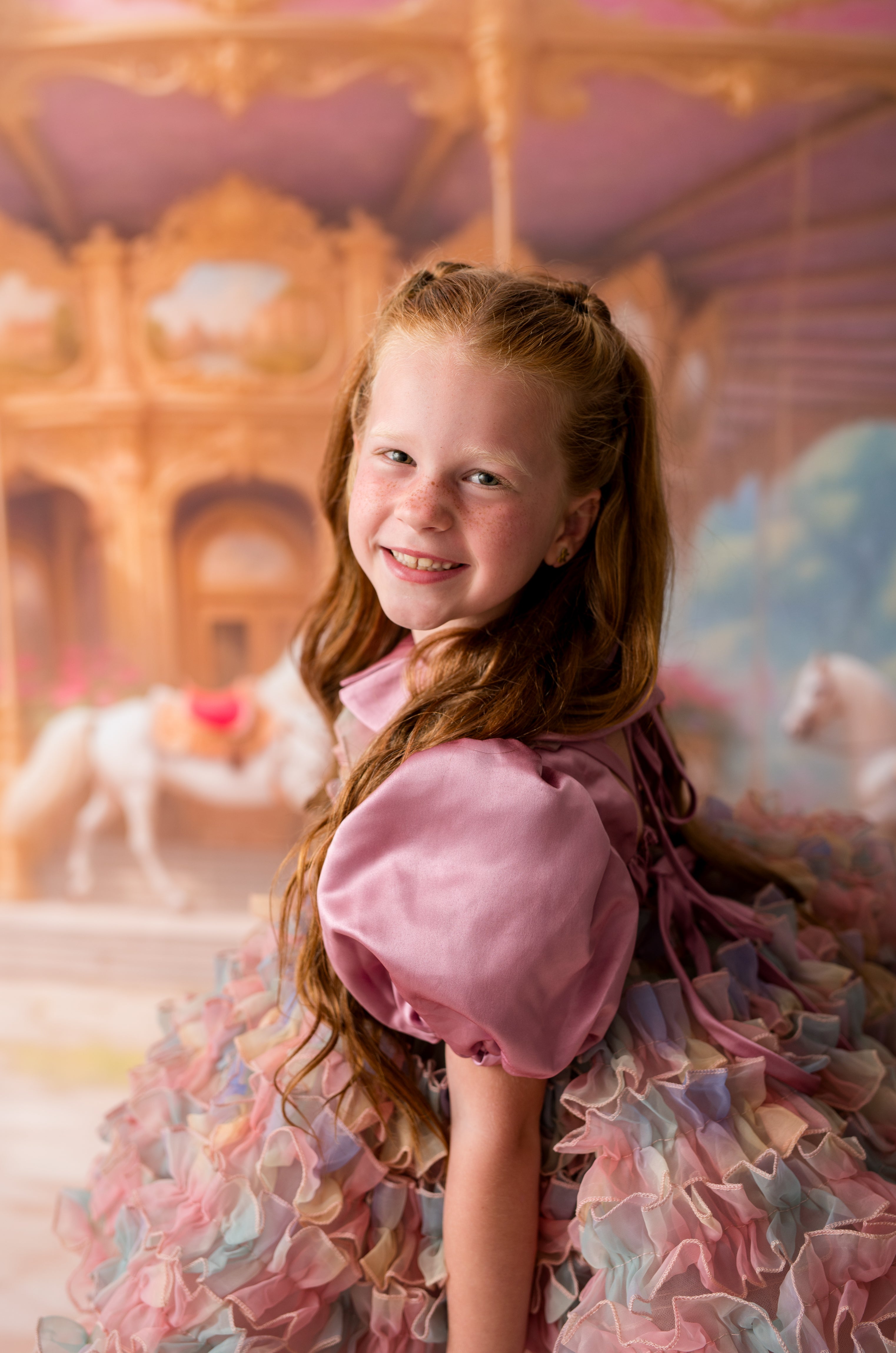 Young girl in a pink dress standing in front of a carousel with horses.