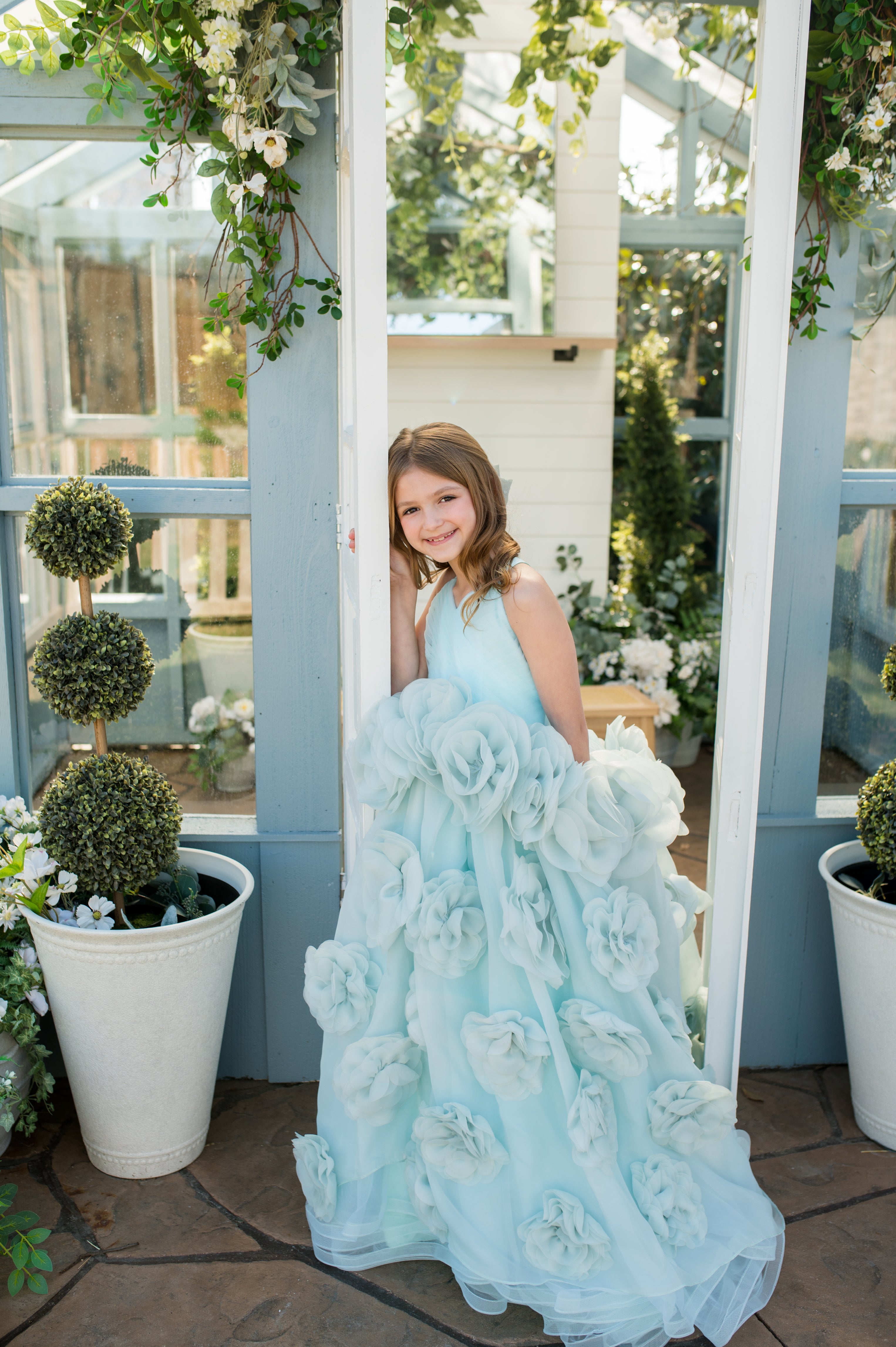 Young girl in a light blue floral dress standing in a greenhouse with plants and flowers around.