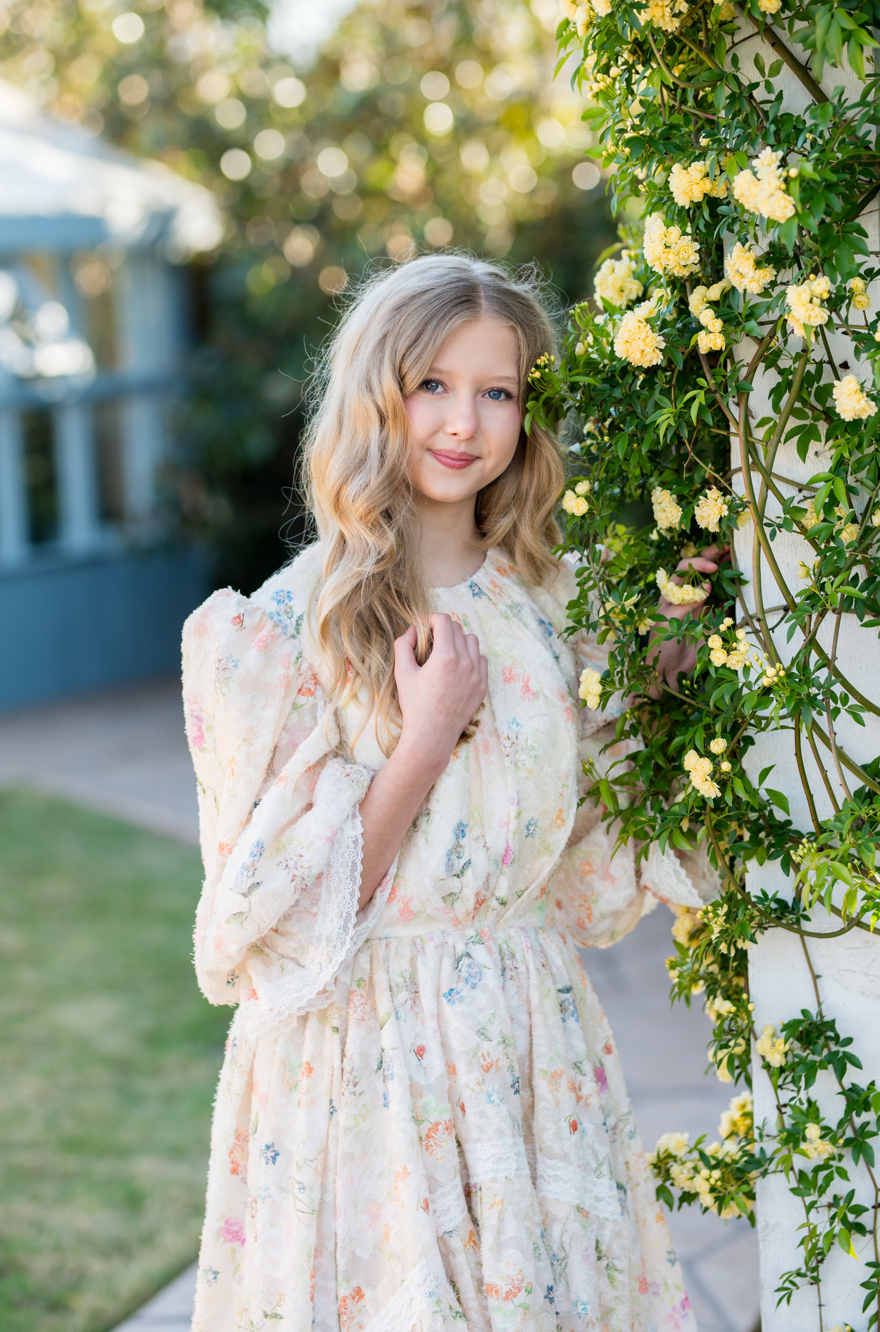 Young girl in a floral dress standing next to a flowering plant outdoors.