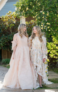 Two women in elegant dresses standing outdoors with greenery and flowers in the background