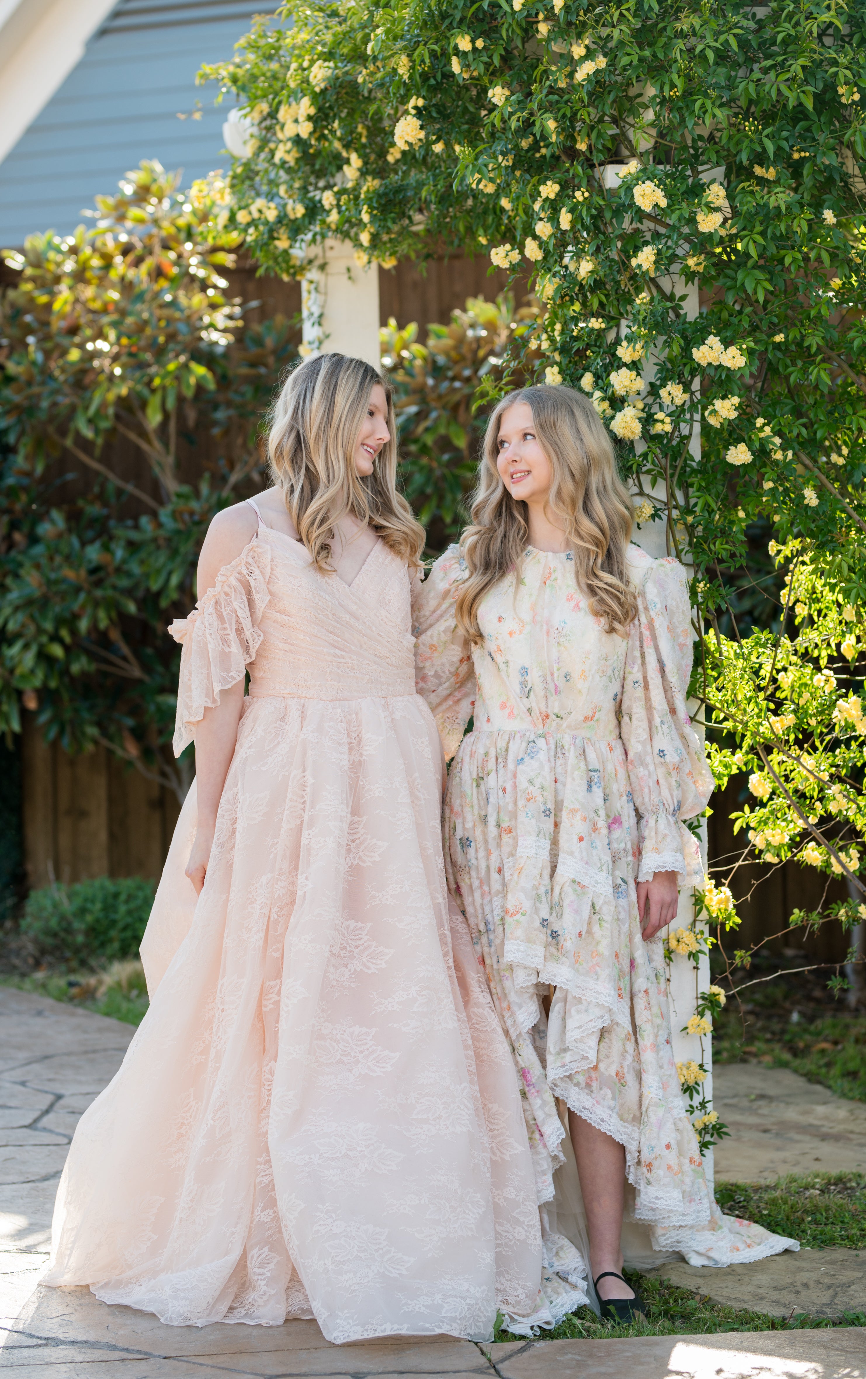 Two women in elegant dresses standing outdoors with greenery and flowers in the background