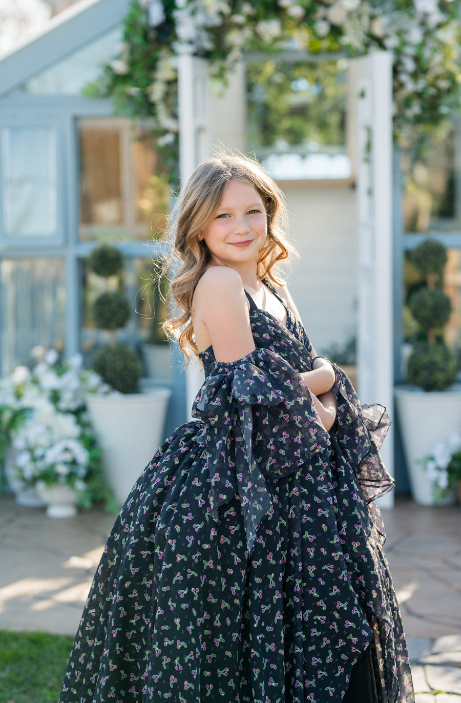 Young girl in a floral dress standing outdoors with decorative flowers and plants in the background.