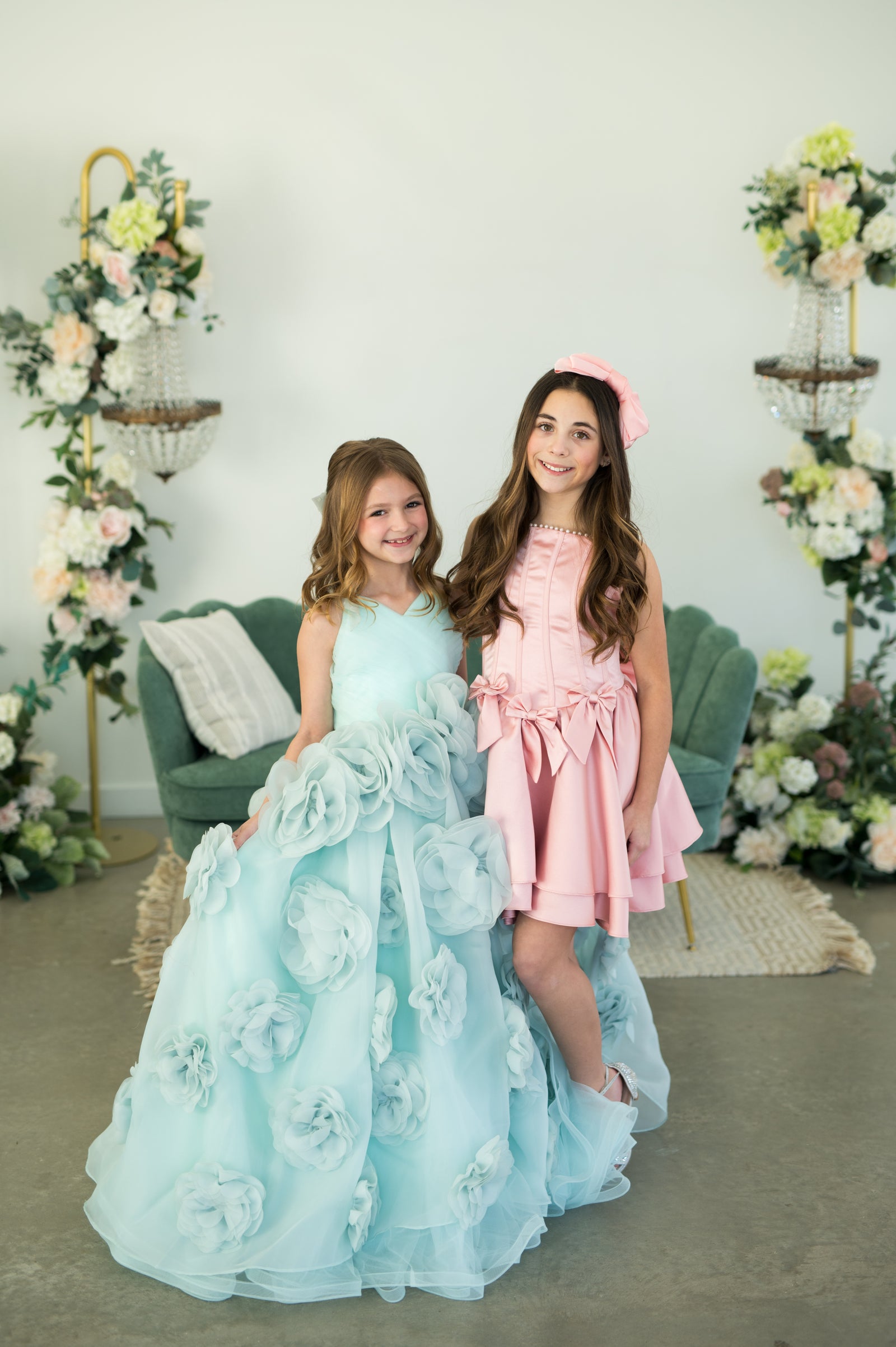 Two young girls in formal dresses standing in a decorated room with floral arrangements.