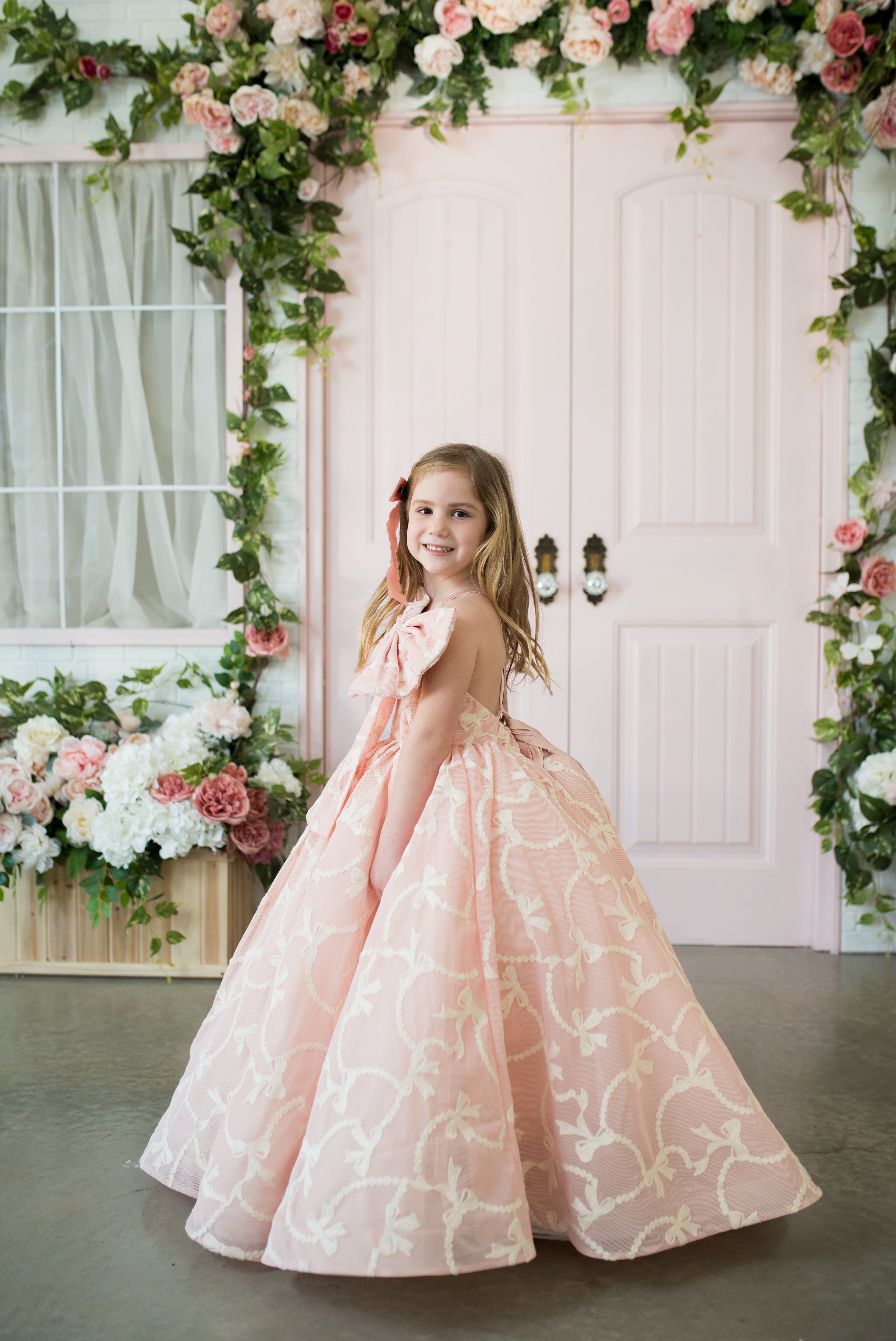 Young girl in a pink floral dress standing in front of a decorated archway with flowers.