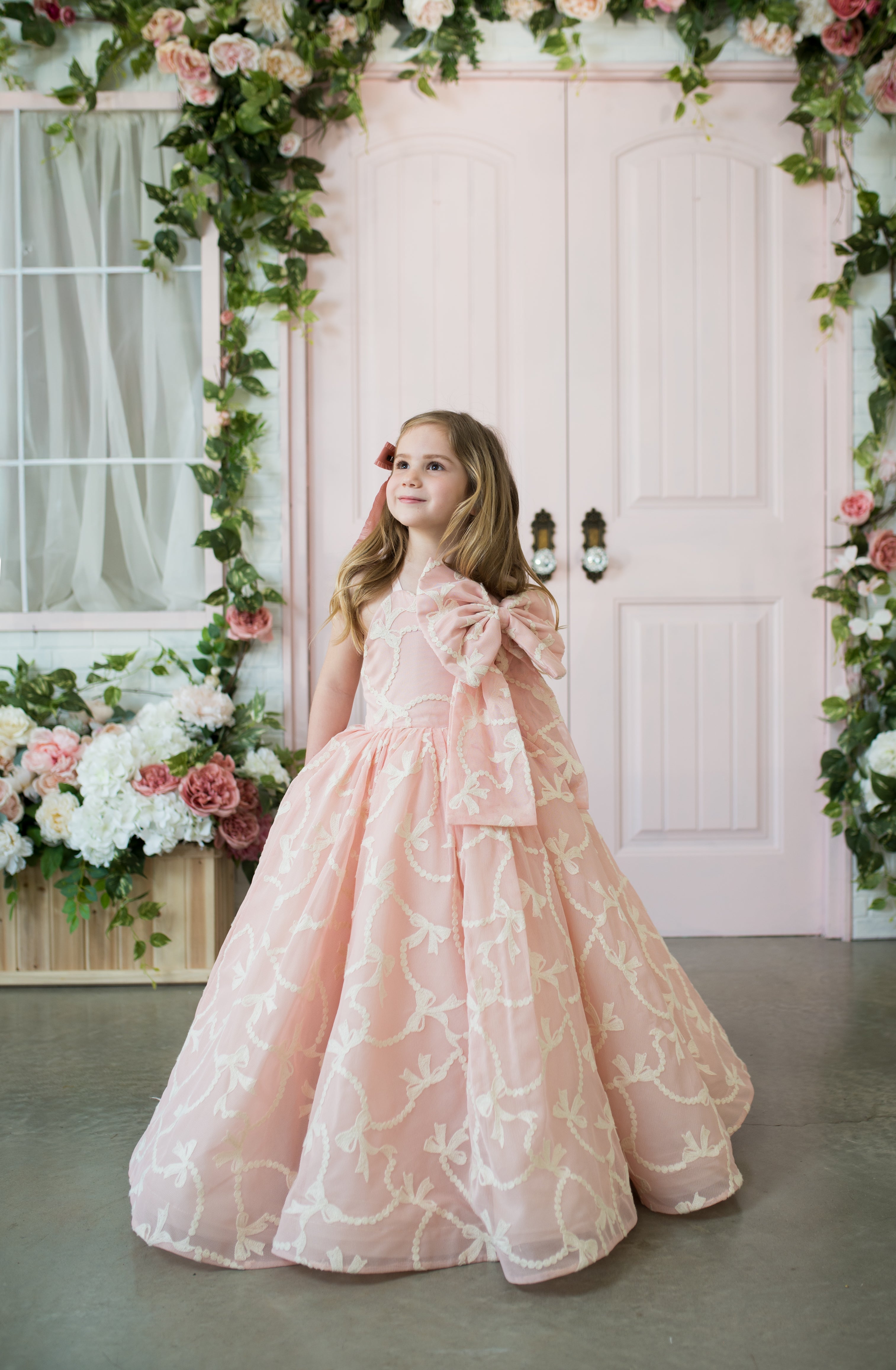 Young girl in a pink floral dress standing in front of a decorated archway with flowers.