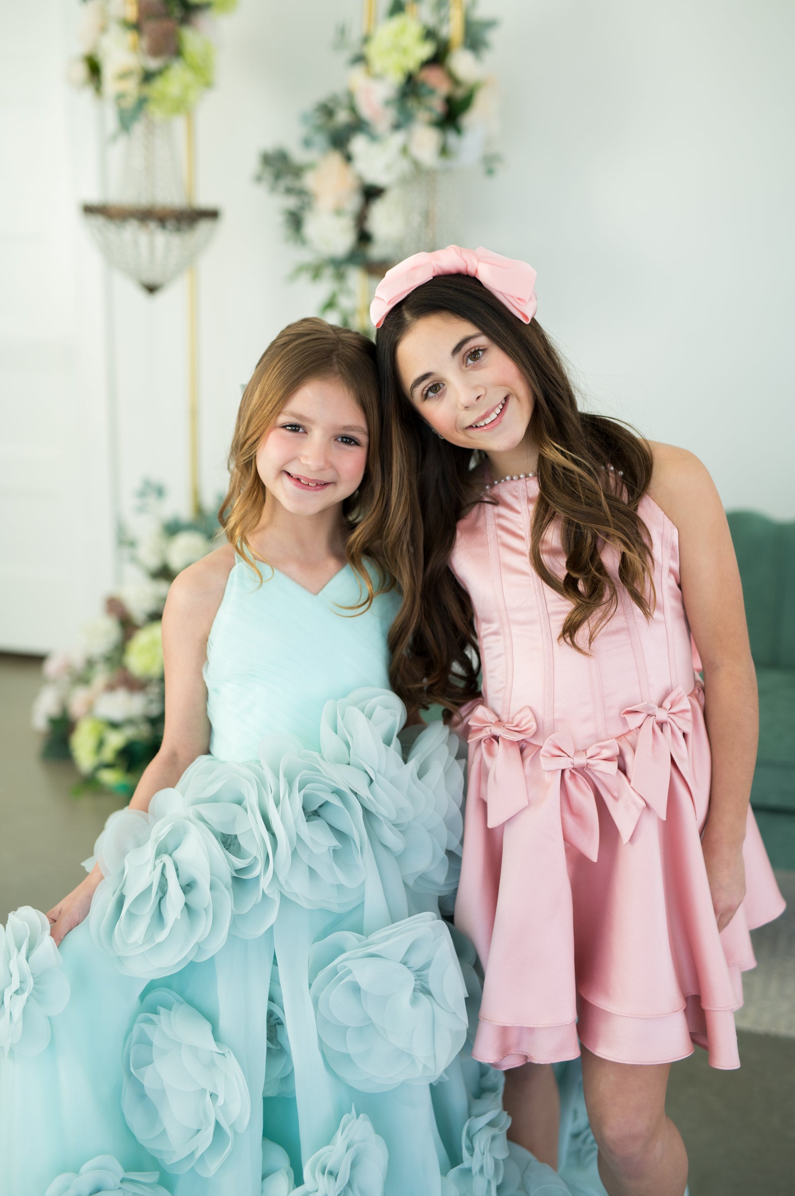 Two young girls in formal dresses standing together with floral decorations in the background.