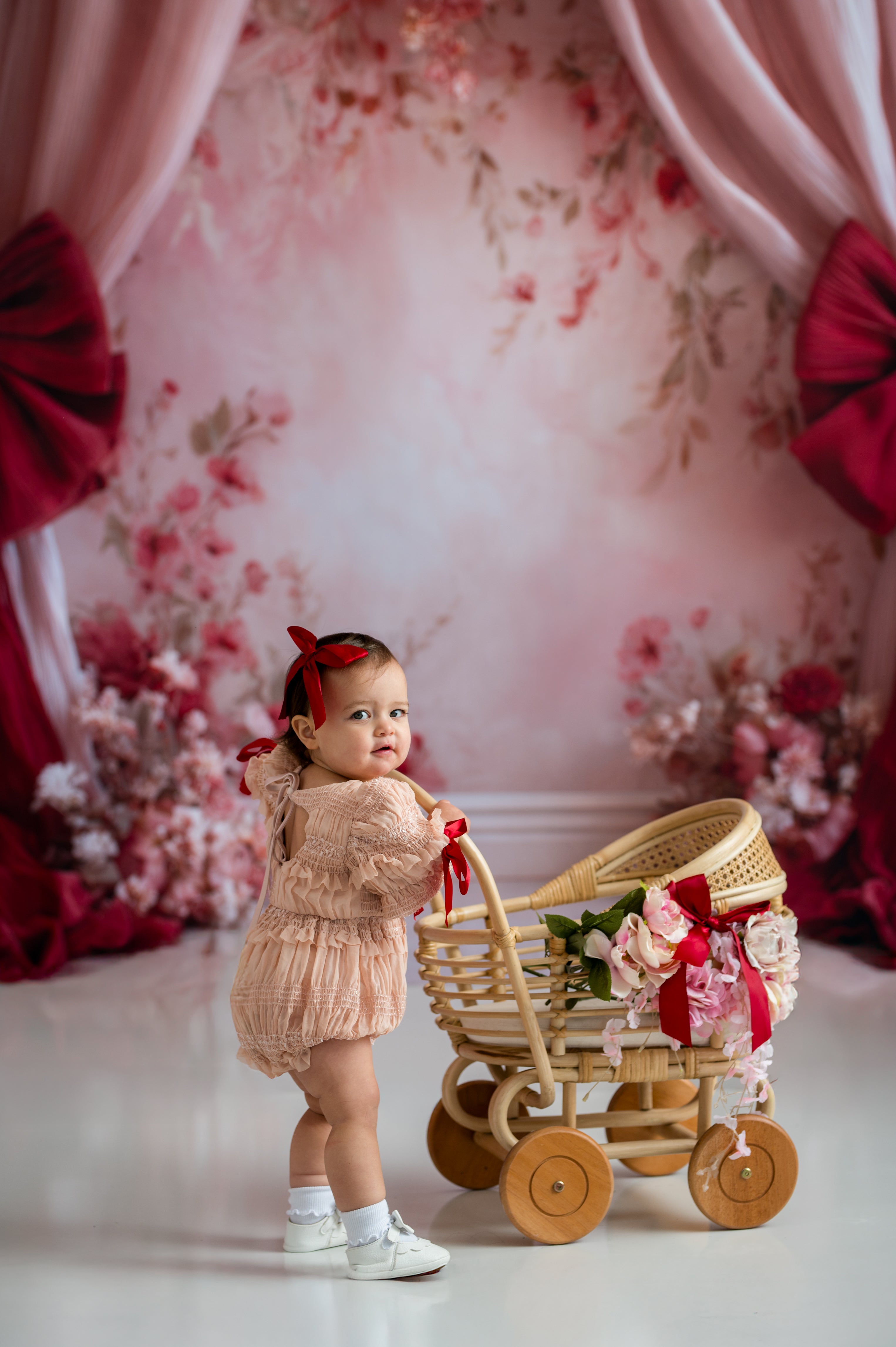 Young child in a pink dress standing next to a toy pram in a decorated room with floral curtains.