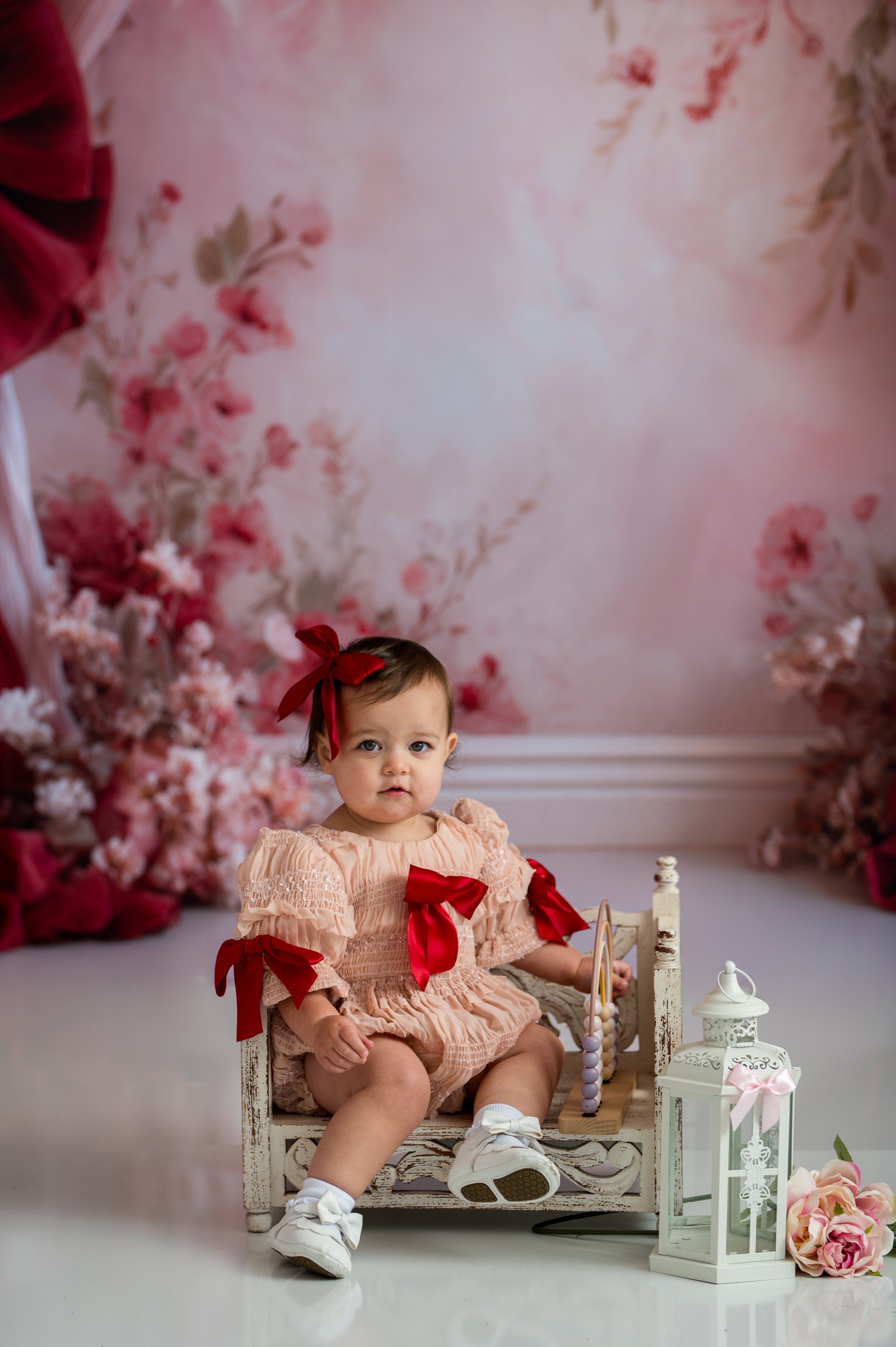 Baby in a pink dress with red bows sitting on a decorative chair against a floral backdrop