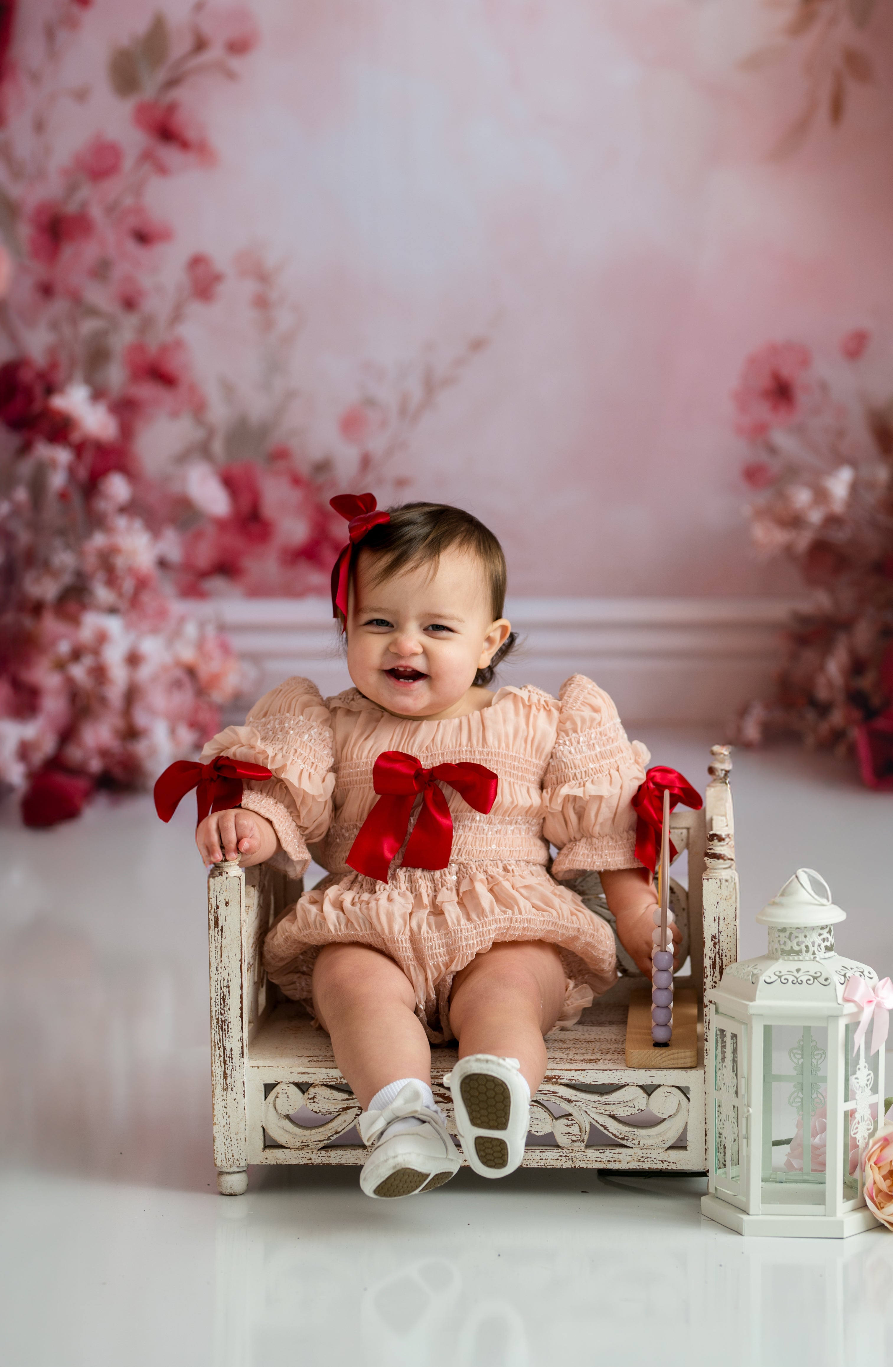 Baby in a pink dress with red bows sitting on a small bed against a floral backdrop