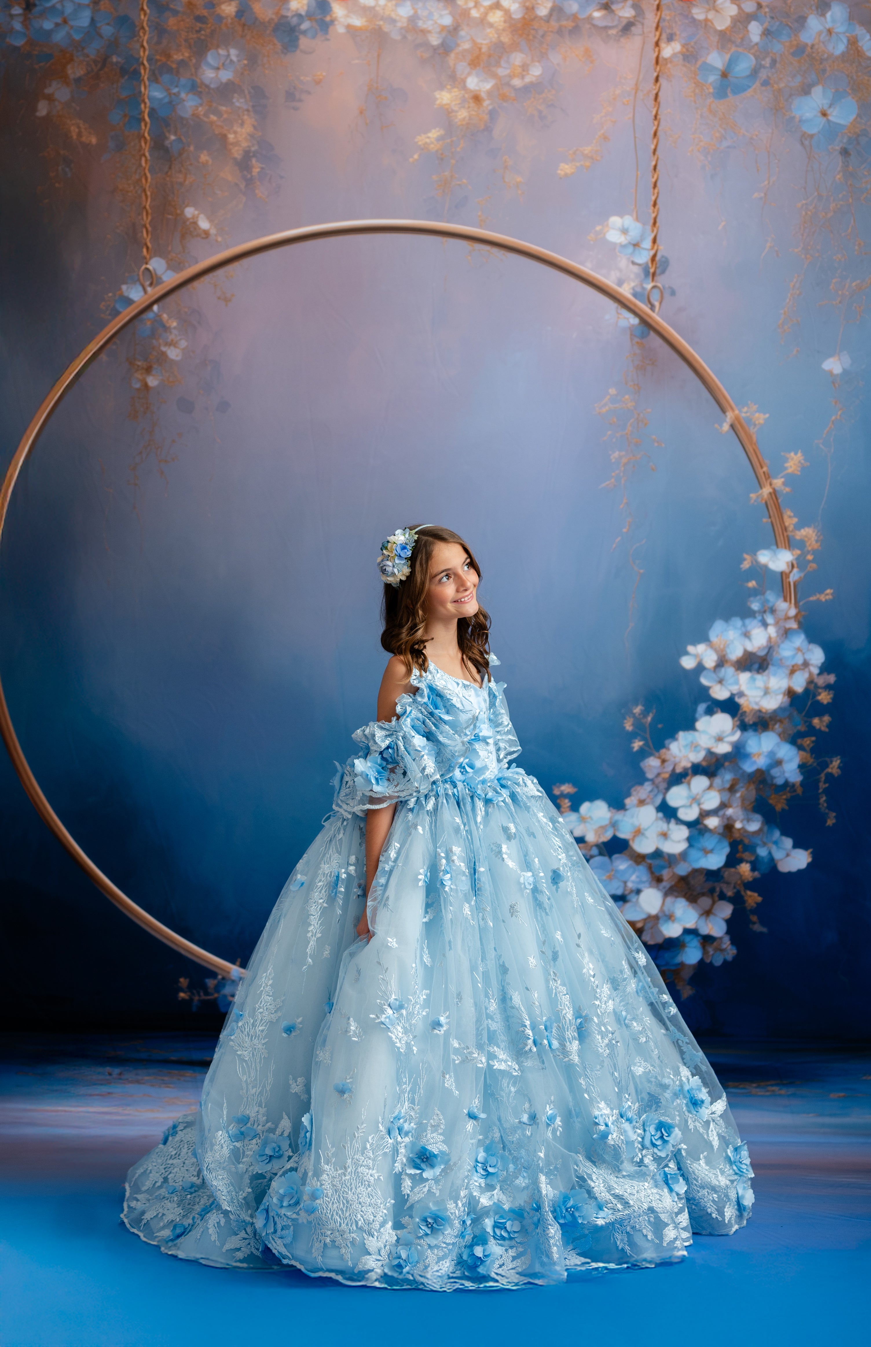 Woman in a blue floral dress standing under a large circular frame with floral decorations against a blue background.