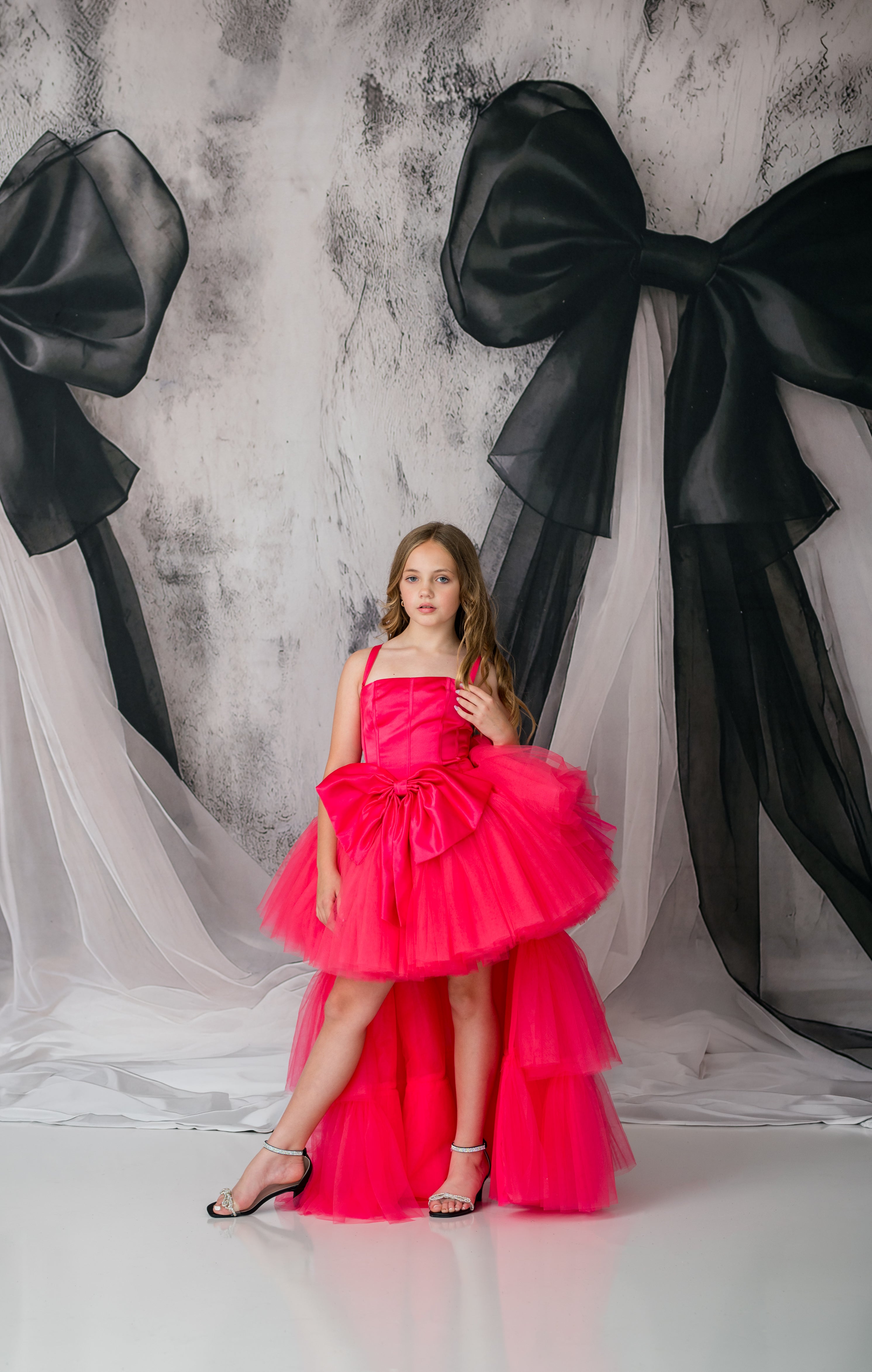 Young girl in a pink dress standing in front of large black bows on a textured wall.