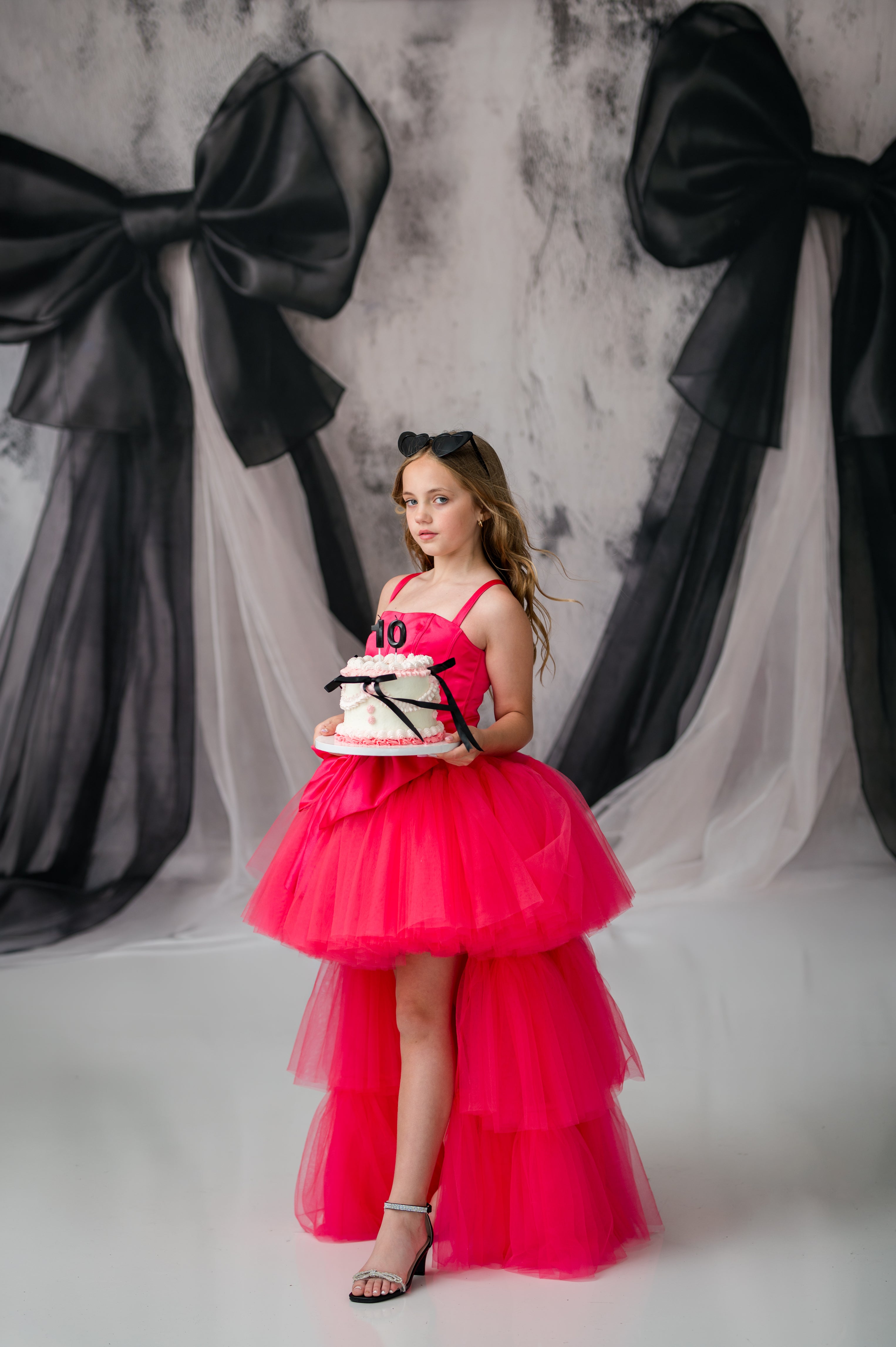Young girl in a pink dress holding a cake with large black bows in the background.