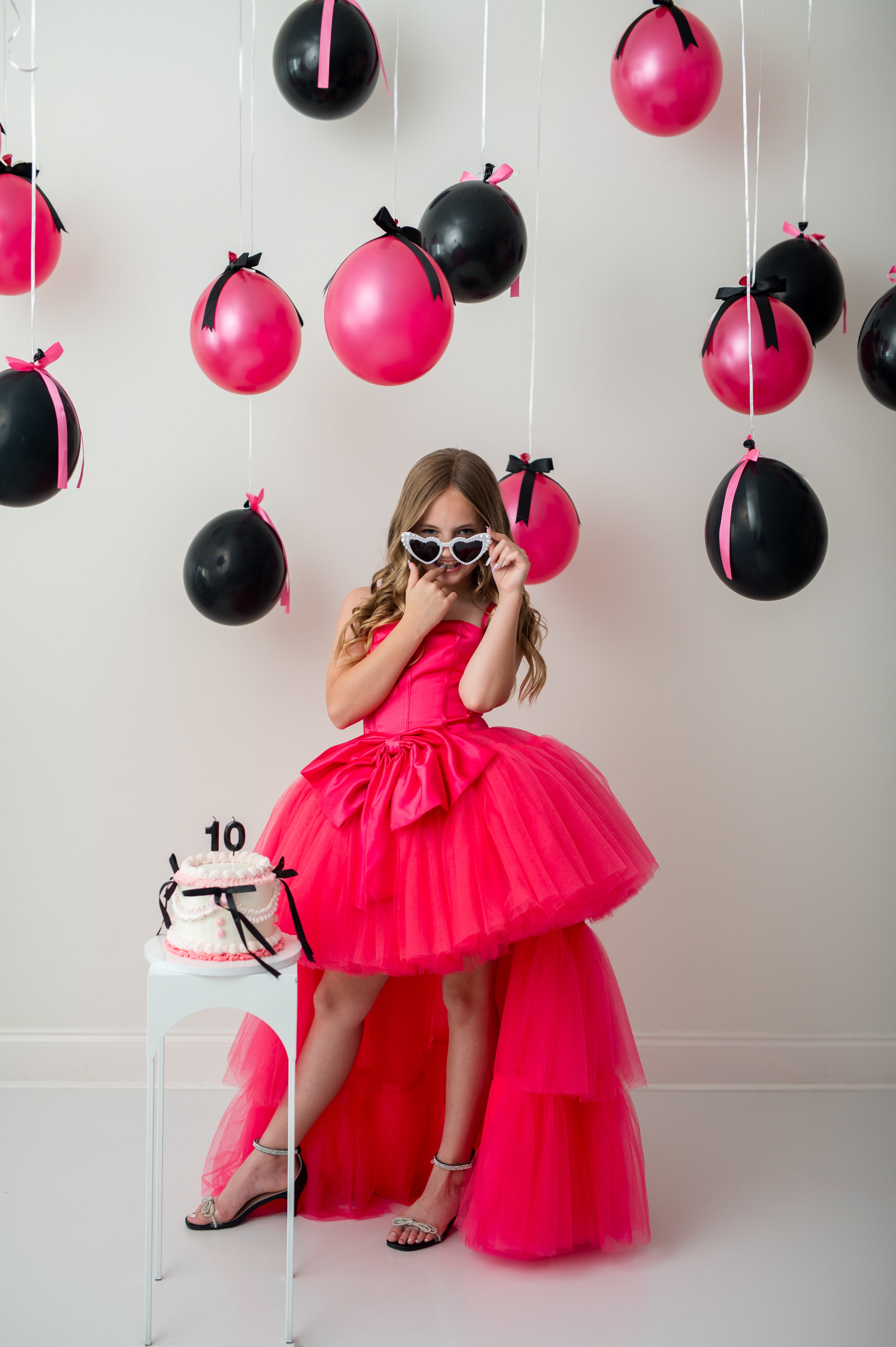 Young girl in a pink dress standing in front of balloons with a cake on a table.