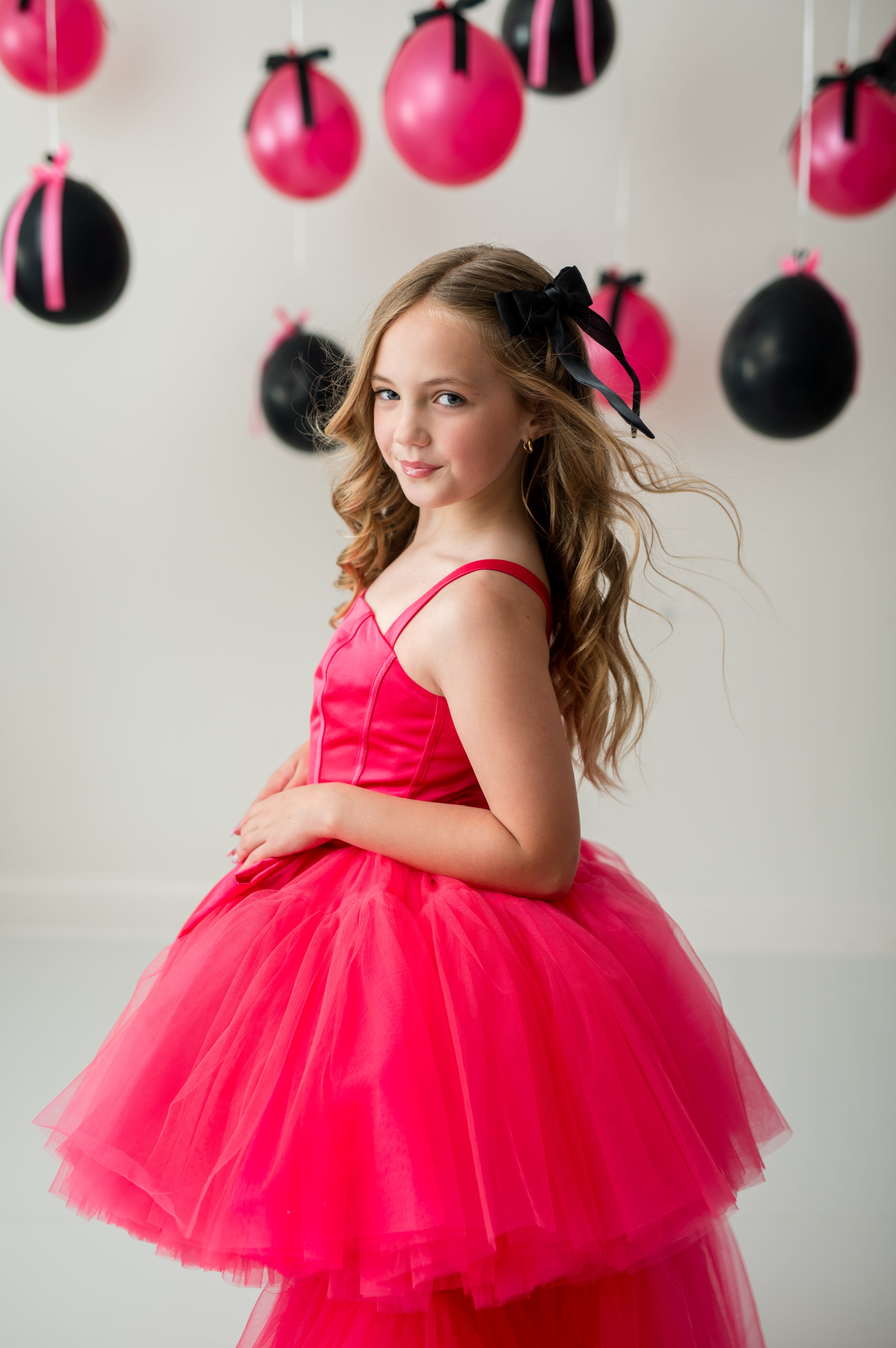 Young girl in a pink dress with black hair bow standing in front of balloons.