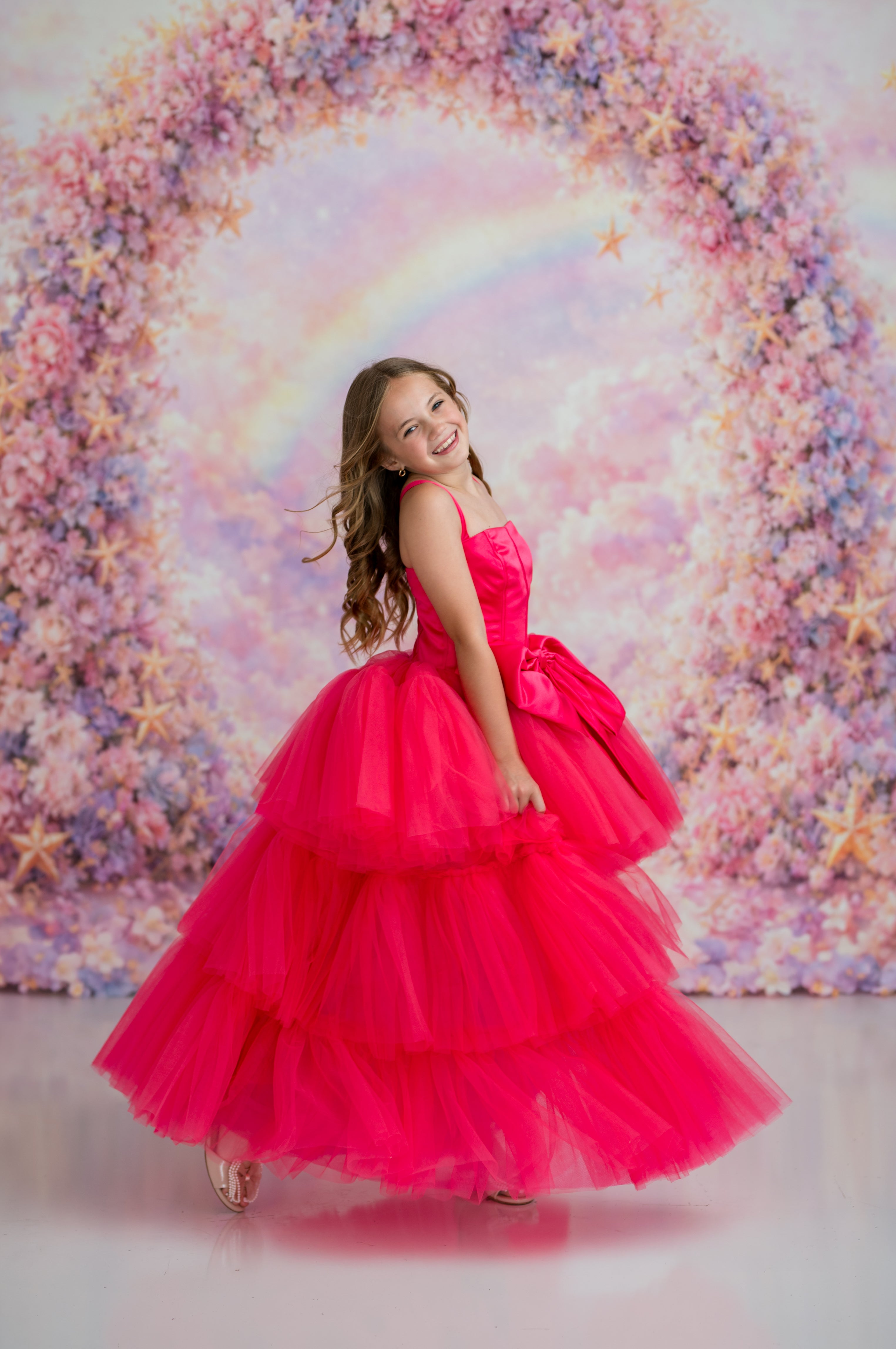 Young girl in a pink dress standing in front of a colorful floral backdrop with a rainbow.