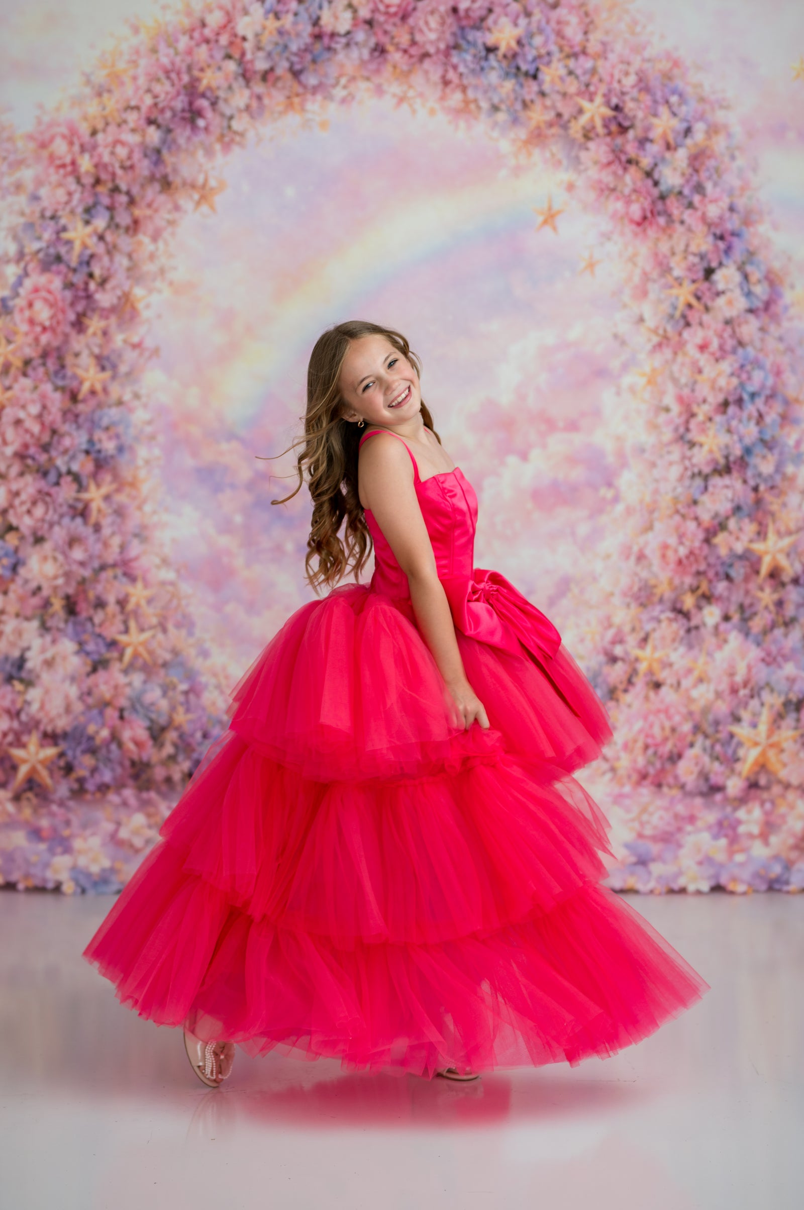 Young girl in a pink dress standing in front of a colorful floral backdrop with a rainbow.