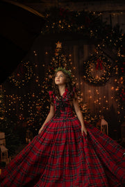A girl dressed in a red and black tartan dress with a black floral lace accent, standing in front of a warmly lit Christmas tree.
