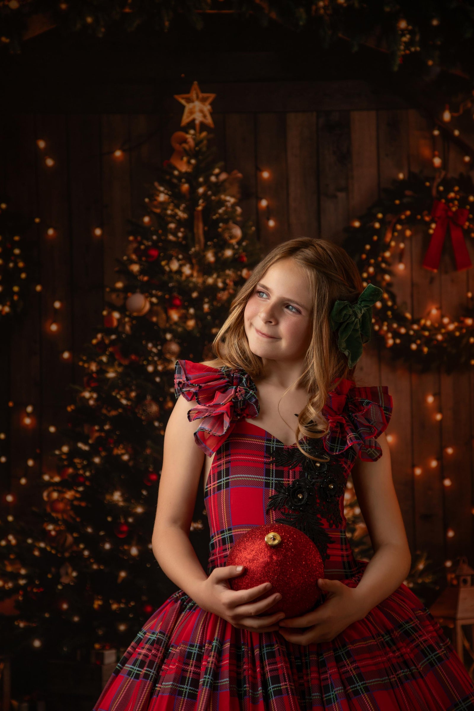 Child smiling in a holiday plaid dress with a decorated Christmas tree and festive lights in the background.

