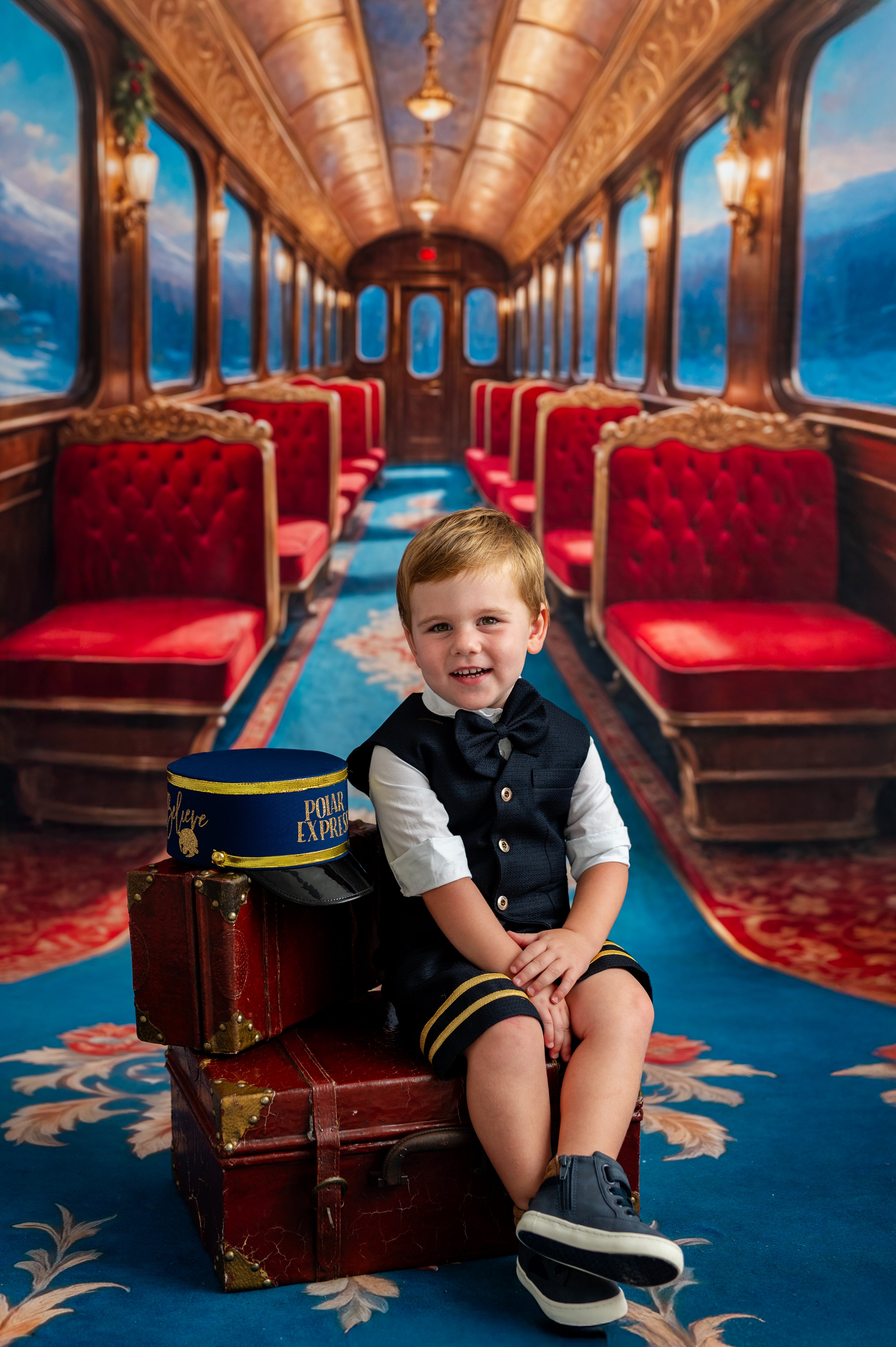 Young boy in a formal outfit sitting on a suitcase inside a luxurious train car with red seats and ornate decor.