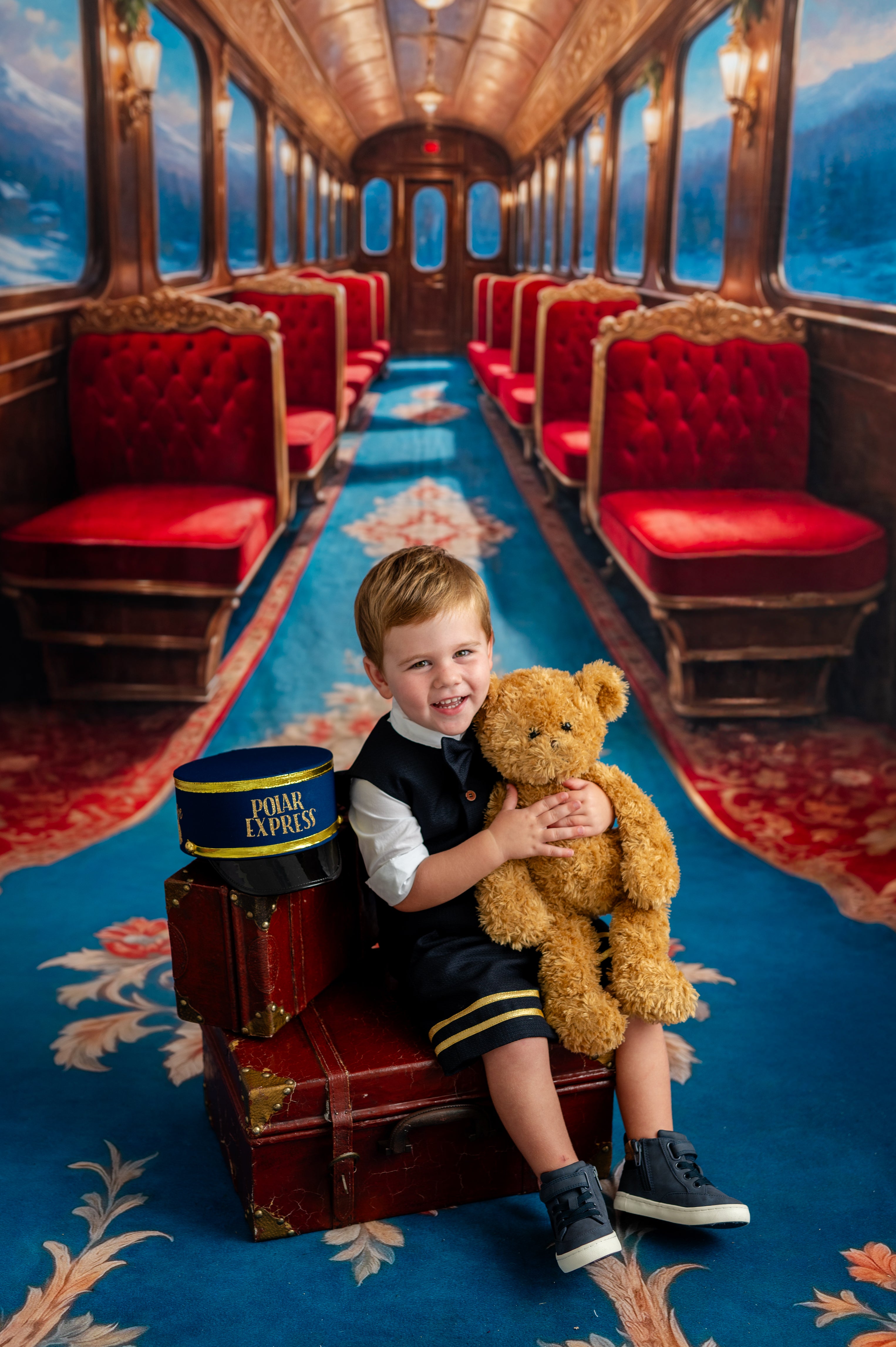 Young boy with a teddy bear sitting on a train car with red seats and a blue carpet.