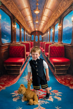 Young boy in a train car with red seats and ornate decor, holding a teddy bear.