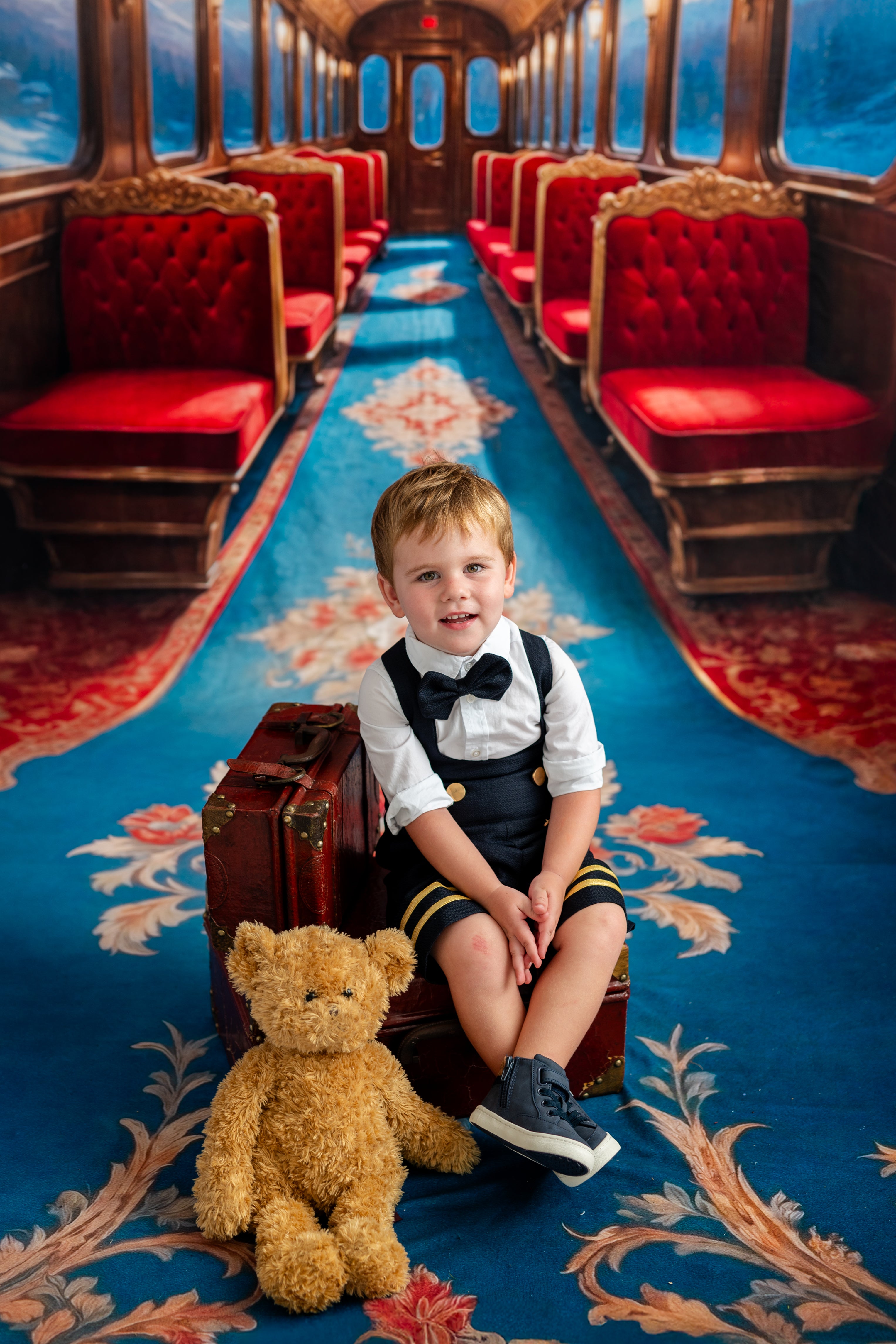 Young boy in formal attire sitting on a suitcase with a teddy bear in a luxurious train car.
