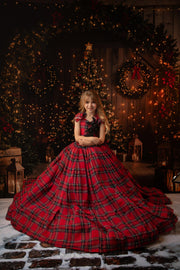 A young girl twirling in a red plaid gown adorned with black lace details, with a warmly lit Christmas tree behind her.
