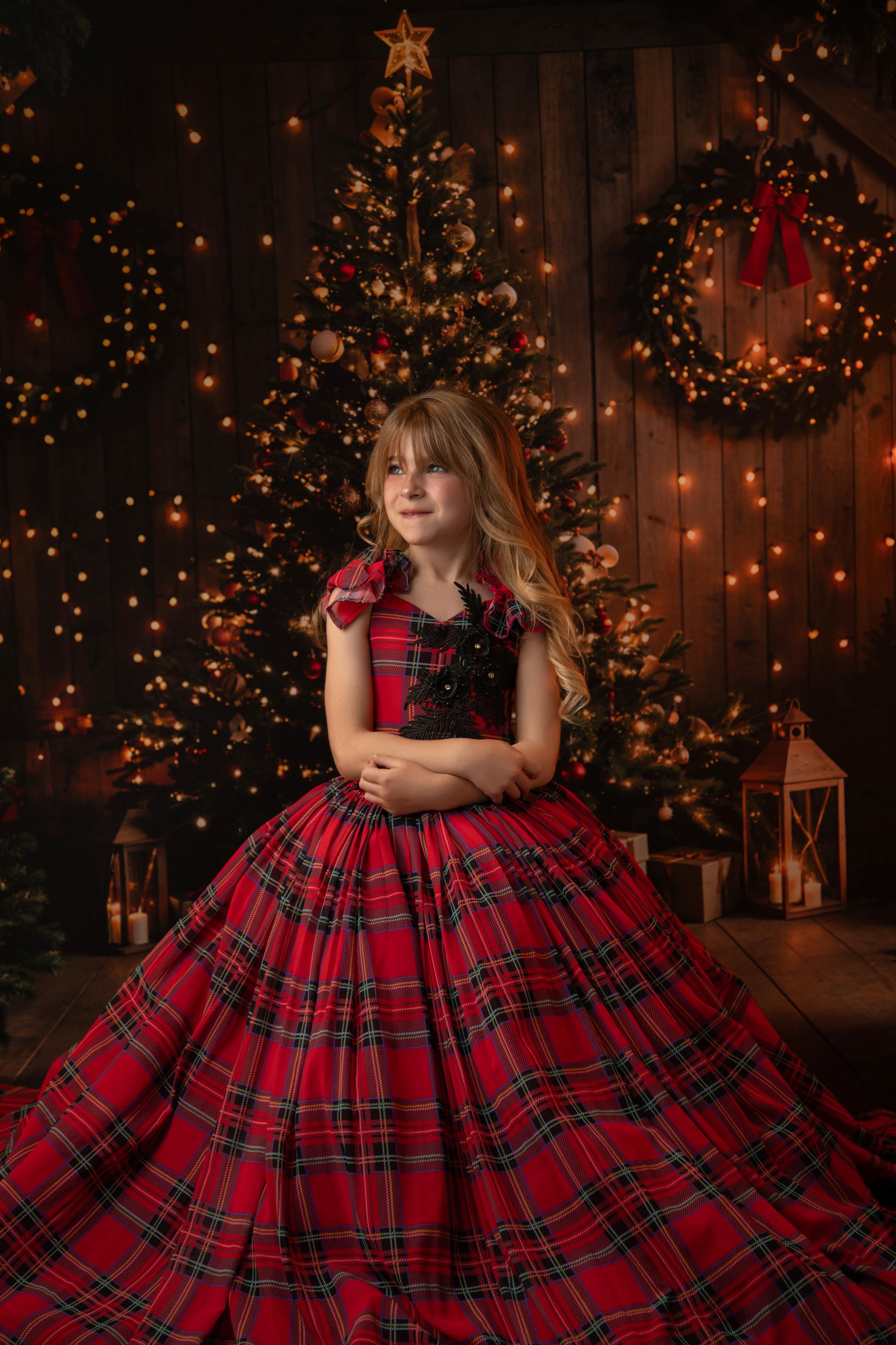 A happy child wearing a festive red tartan dress with floral black appliqué, posing in front of a glowing Christmas scene.
