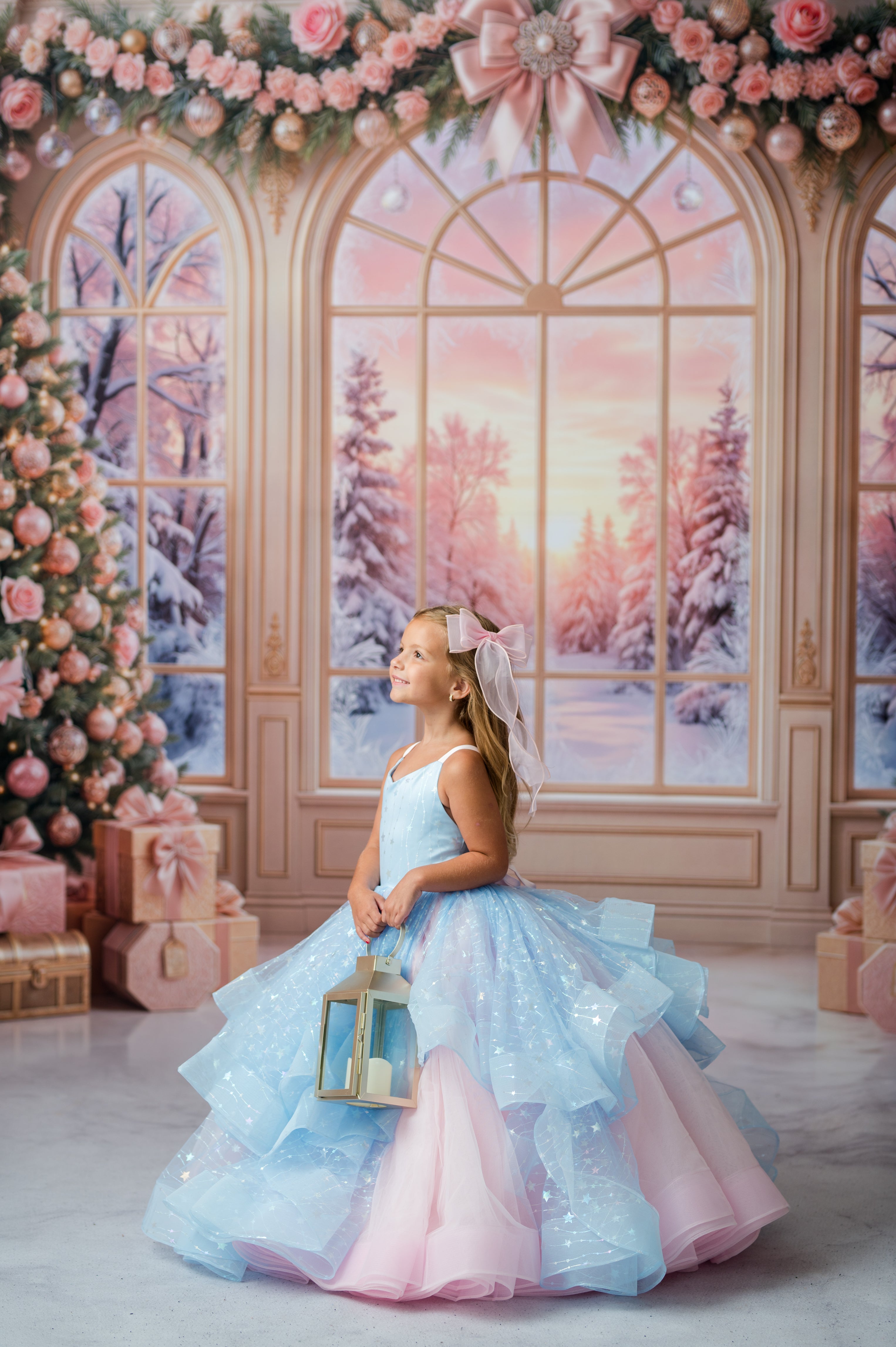 A young girl stands in a decorated indoor studio scene wearing a voluminous ball gown with a light blue and pink color-blocked design and sparkly details. She holds a small lantern, with a pink bow in her hair.