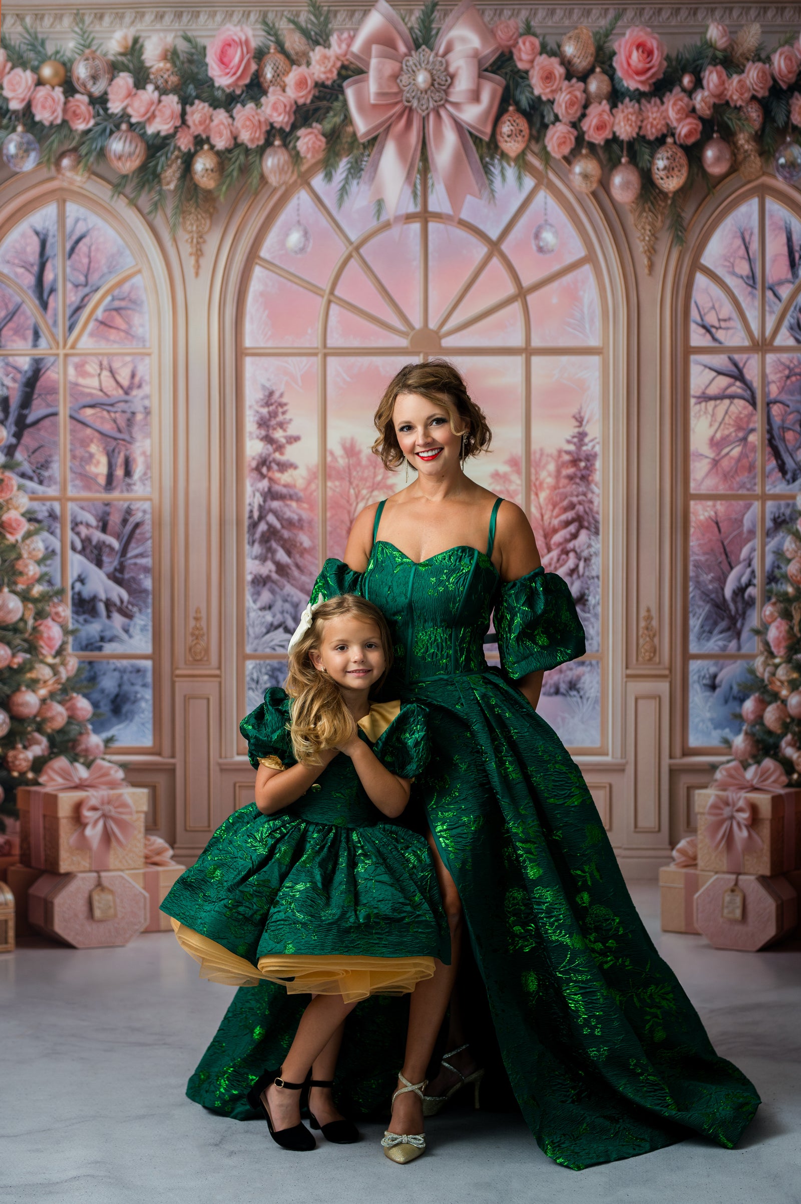A woman and a young girl wearing matching emerald green ballgown-style dresses pose together in front of a festive holiday studio backdrop with pink windows, floral garlands, trees, and gift boxes.