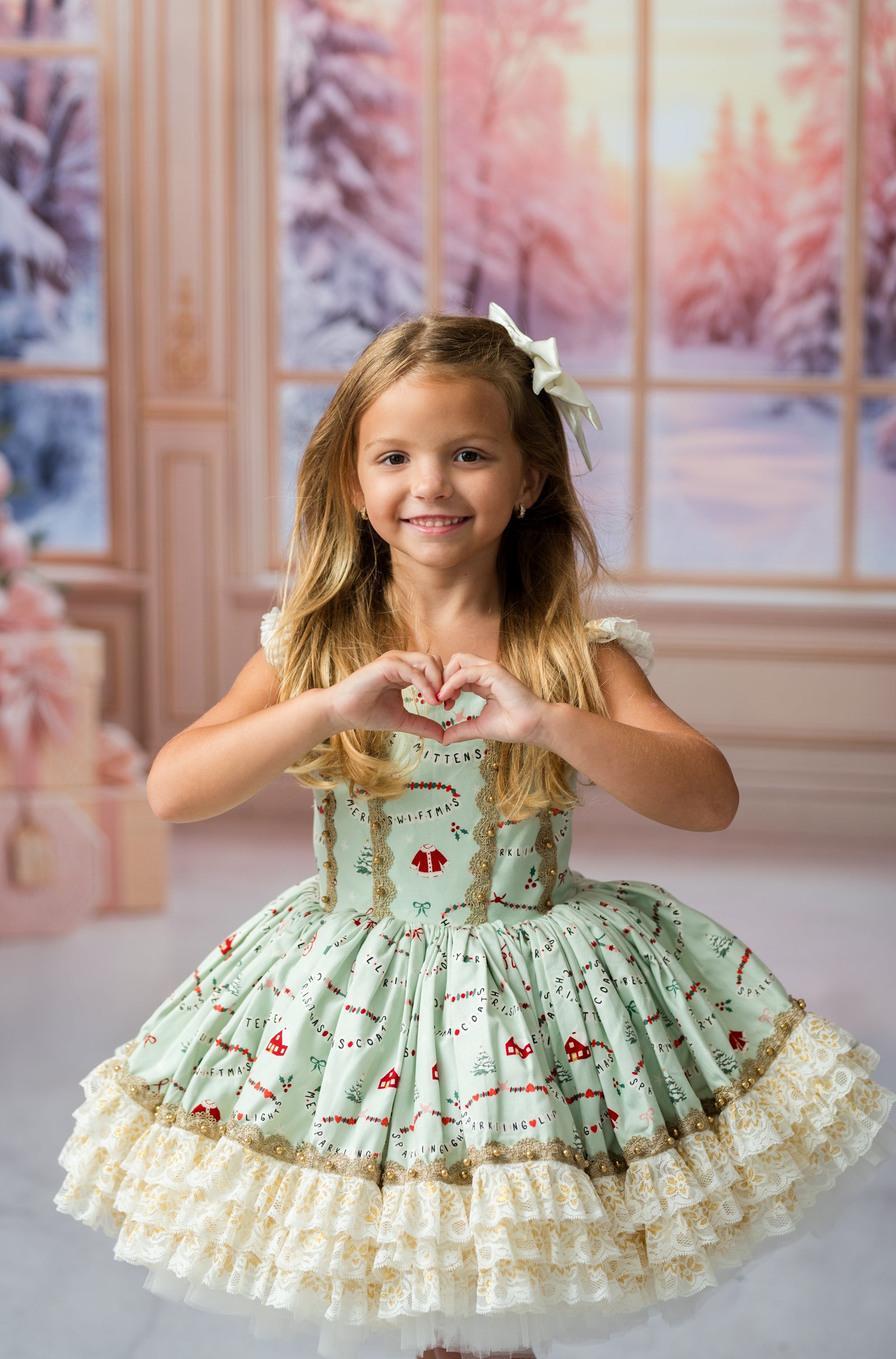 A young girl with long blonde hair wearing a light green and white patterned dress with a white bow in her hair, posing and making a heart shape with her hands. The background is a soft, warm-toned indoor scene with a window showing a snowy forest.