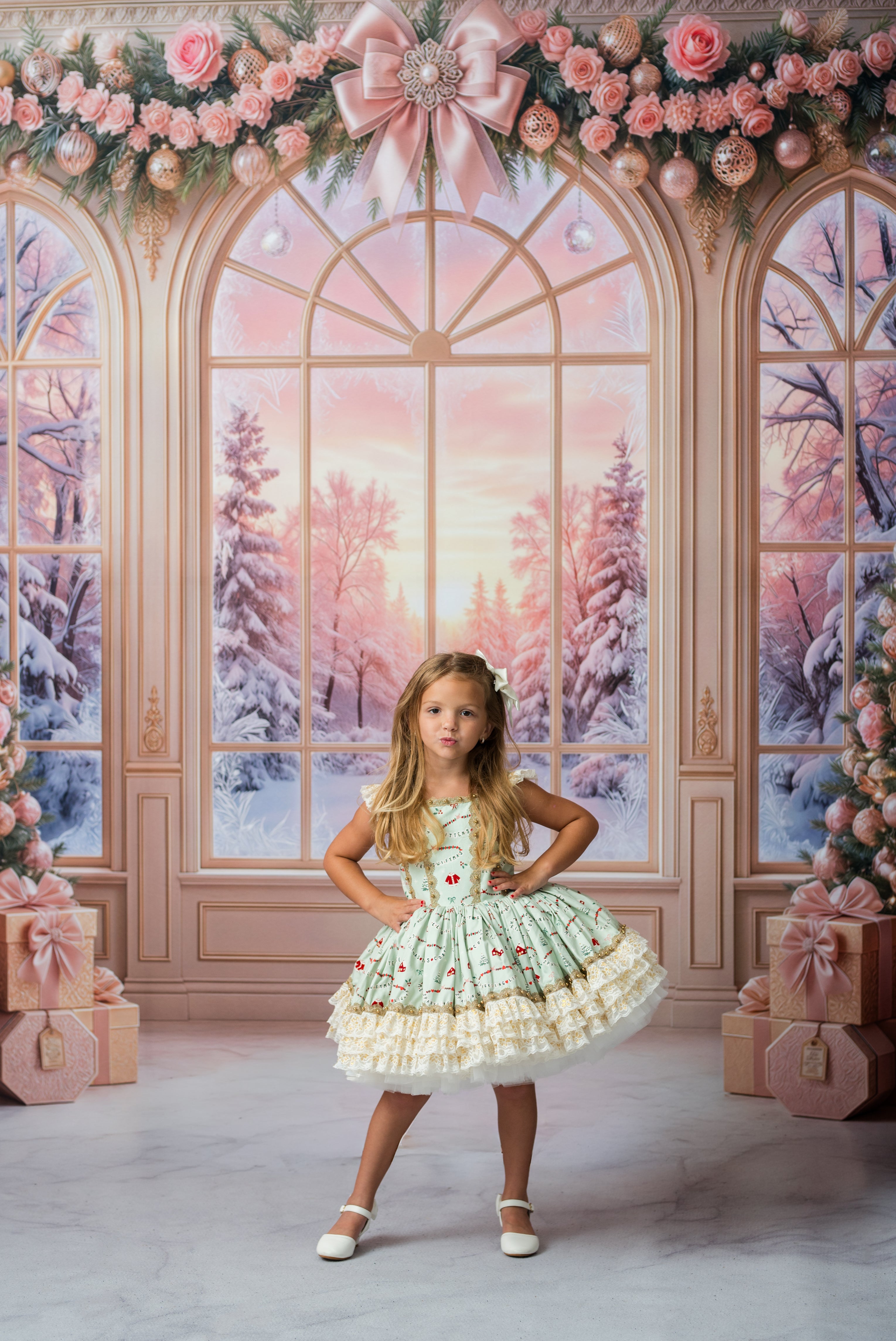 A young girl stands in a festive holiday-themed studio setting with large arched windows showing a snowy winter forest scene. She is wearing a light green and white ruffled dress with small red and pink floral details.